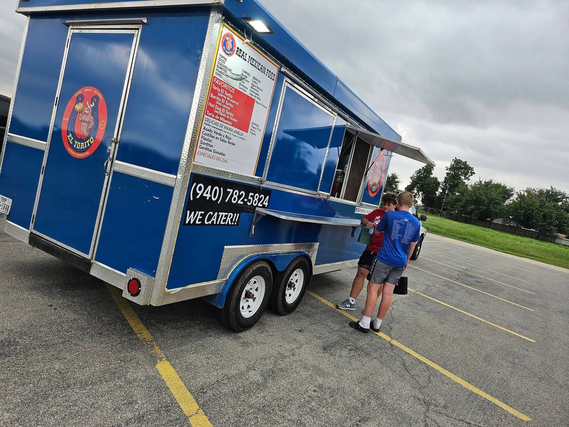 Blue food truck with two people at the serving window in a parking lot.