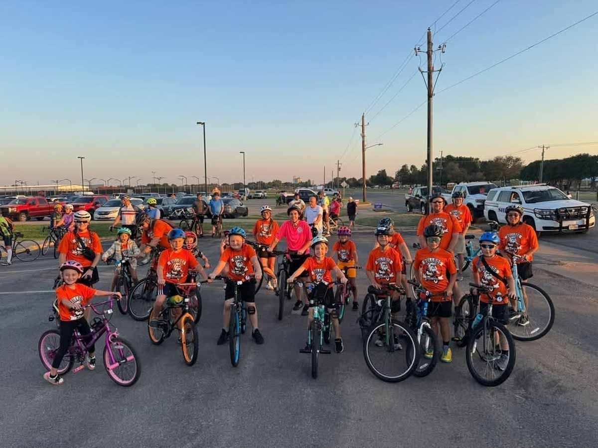 Group of cyclists in orange shirts poses on bikes in parking lot. Cars and buildings in background.