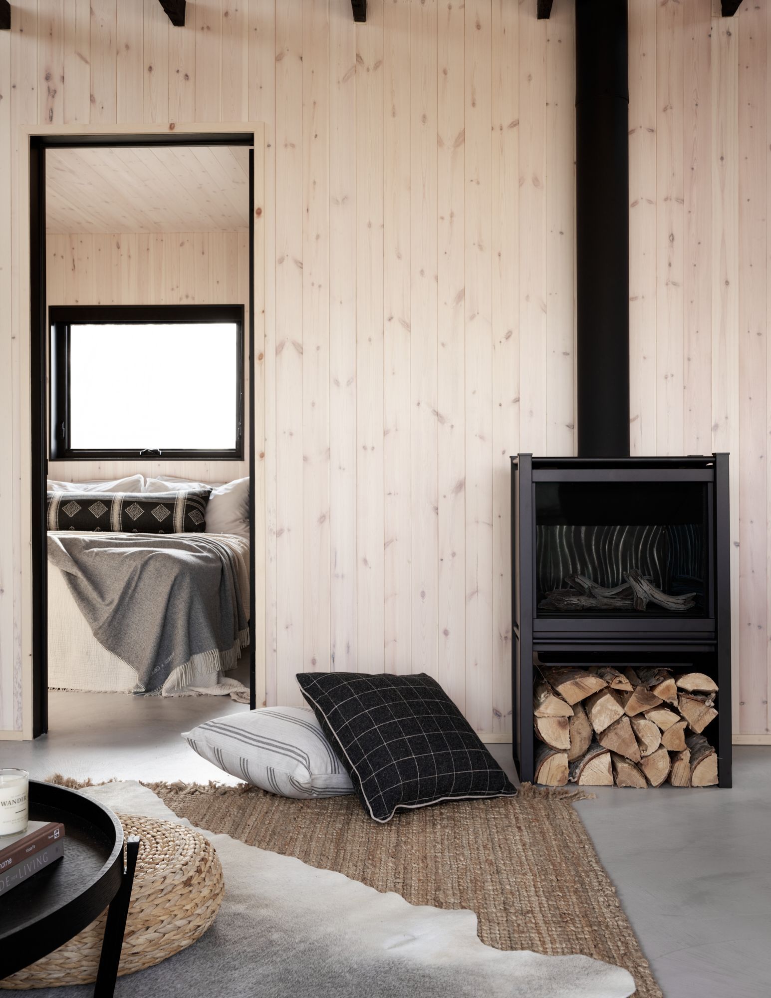 Modern cabin interior with fireplace, stacked logs, and open doorway to bedroom.