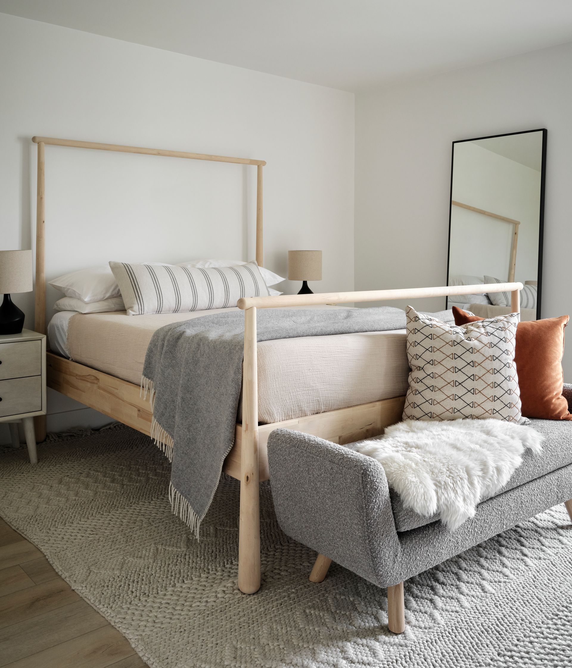 Bedroom with light wood bed, bench, and rug. Cream, gray, and rust-colored accents. Mirror on wall.