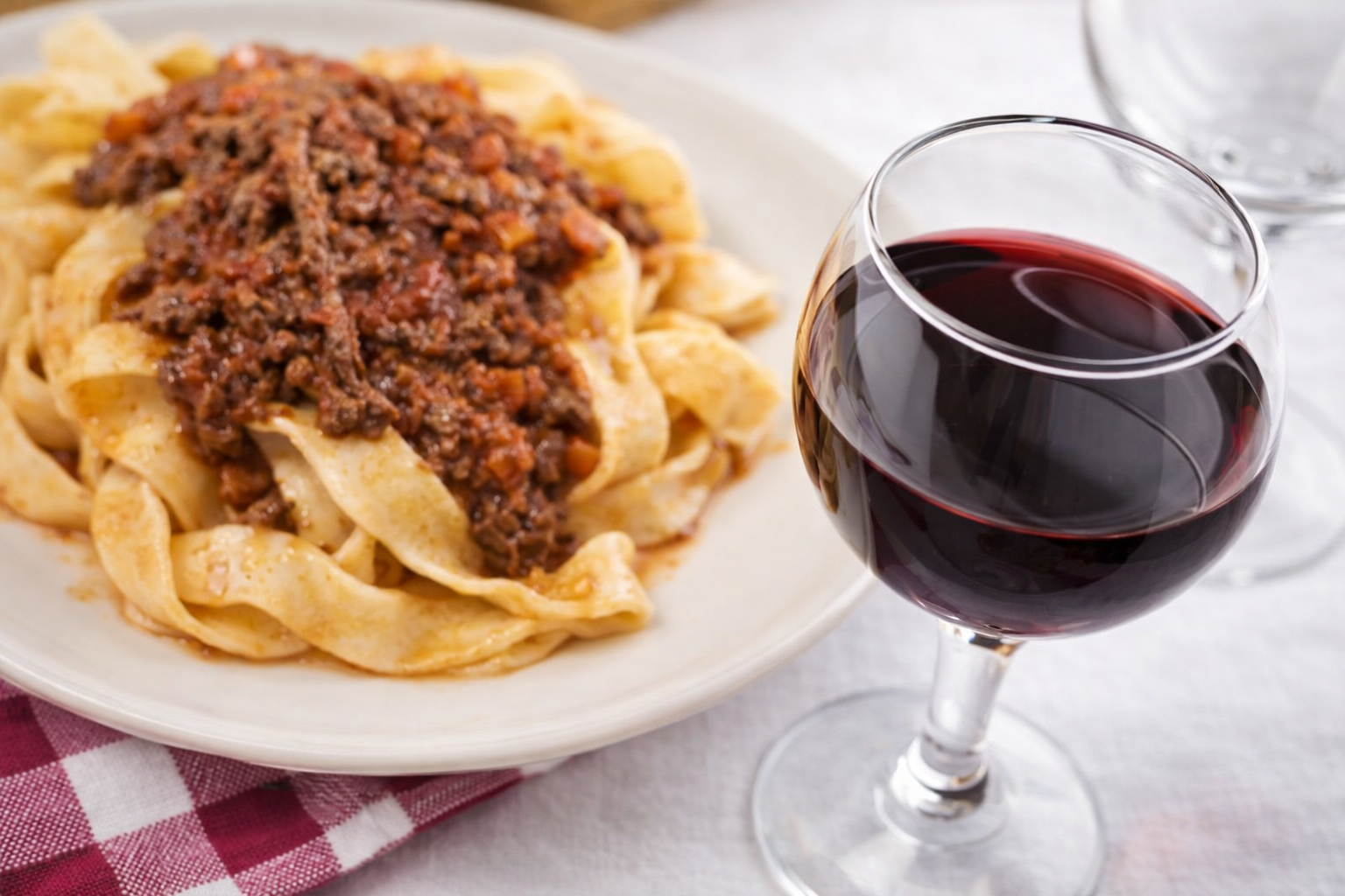 Pasta al ragù, servita accanto a un bicchiere di vino rosso, su un tavolo con un tovagliolo a quadretti rossi.