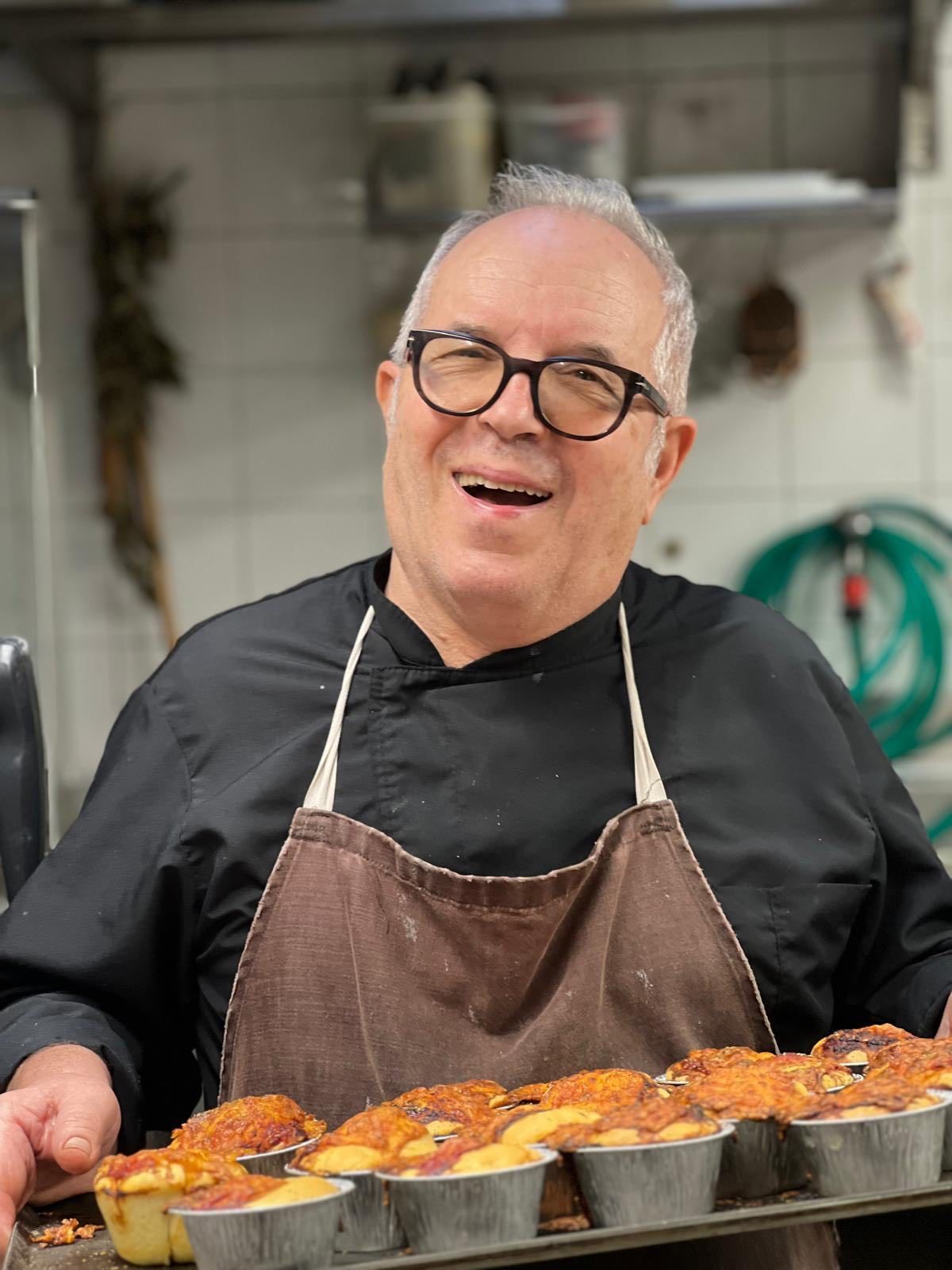 Un uomo con gli occhiali tiene in mano un vassoio di prodotti da forno, sorridendo in cucina.