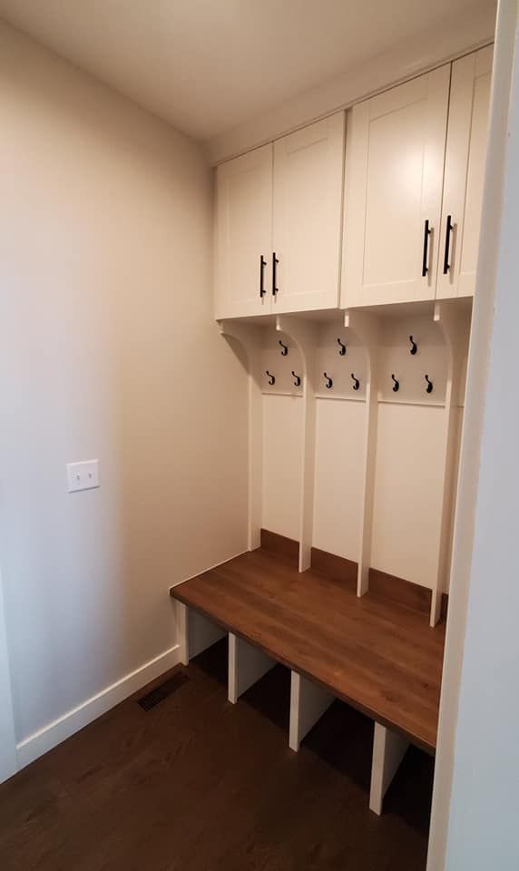 White built-in entryway locker with wood bench, storage cabinets above, and hooks for coats on a dark wood floor.