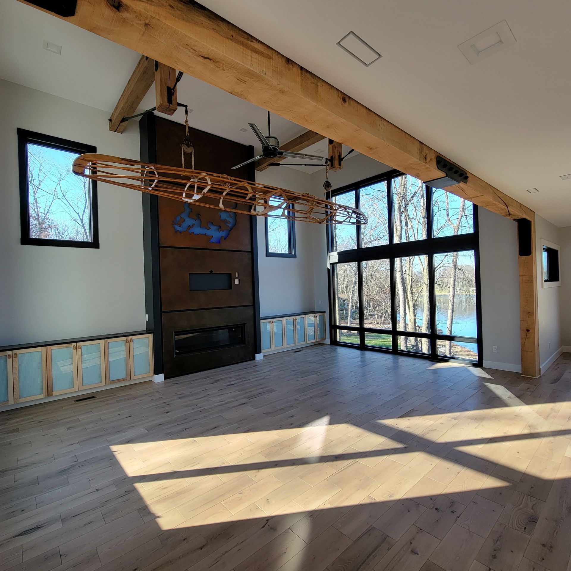 A modern living room with hardwood floors, a large central fireplace, ceiling beams, and tall windows overlooking a lake.