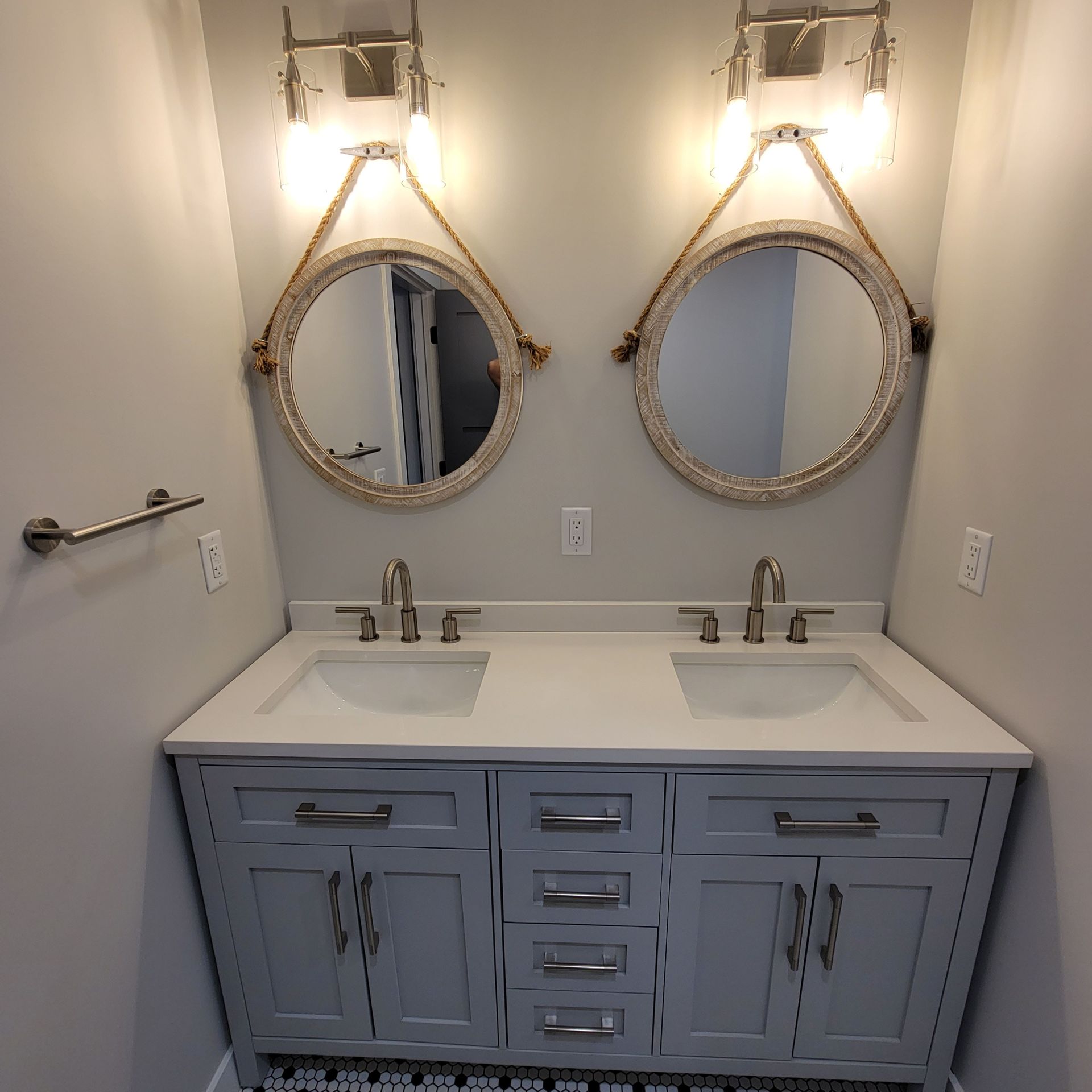 A double-sink bathroom vanity with light gray cabinets, white countertops, rope-framed mirrors, and dual light fixtures.