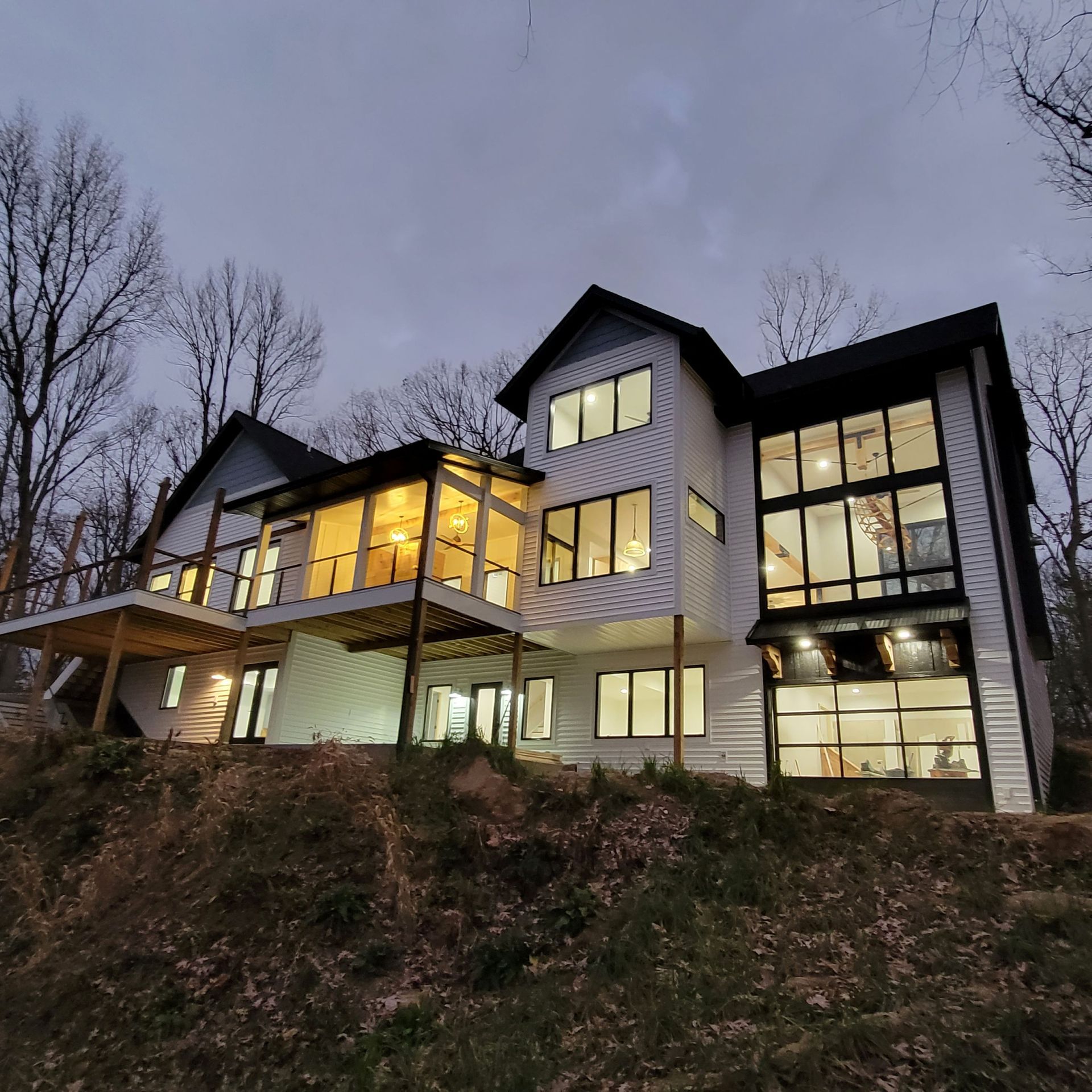 A modern, multi-story house with white siding and large windows, lit from within, set on a wooded hillside at dusk.