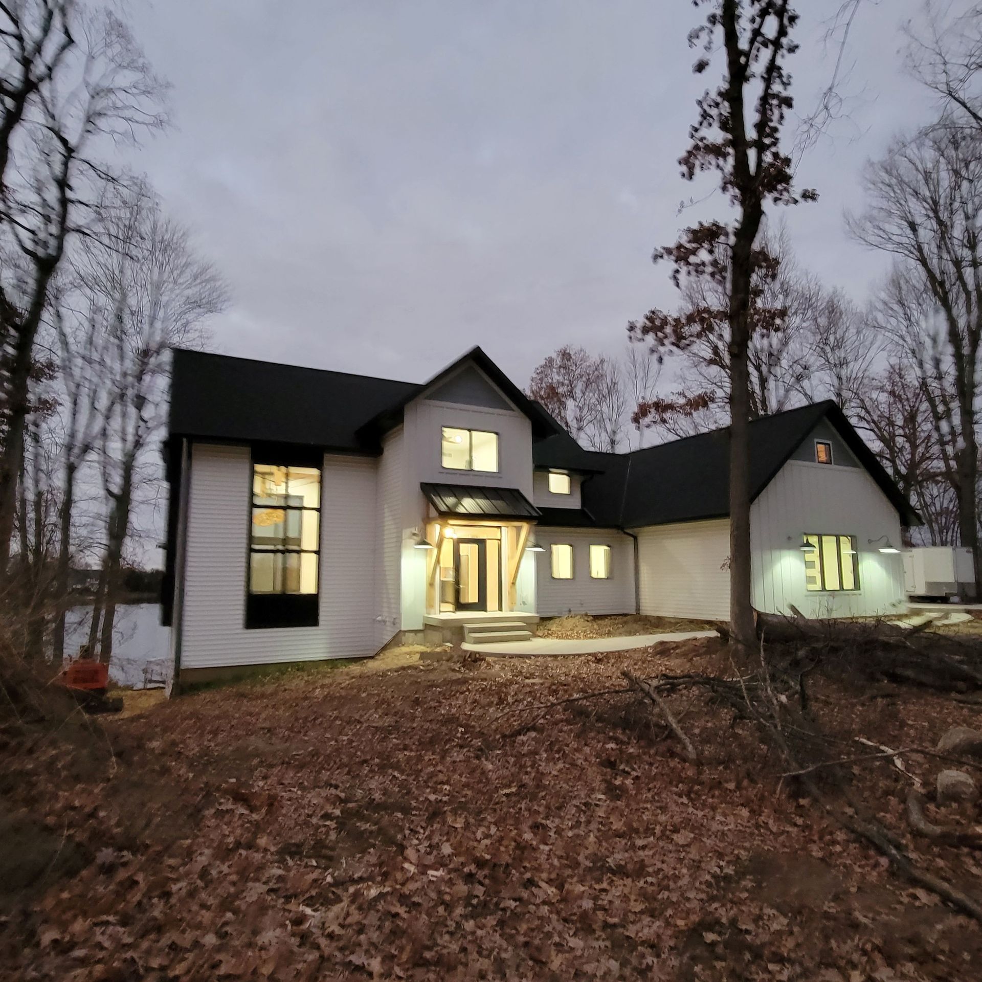 A modern white house with a dark roof and large windows sits at dusk among bare trees and fallen leaves.