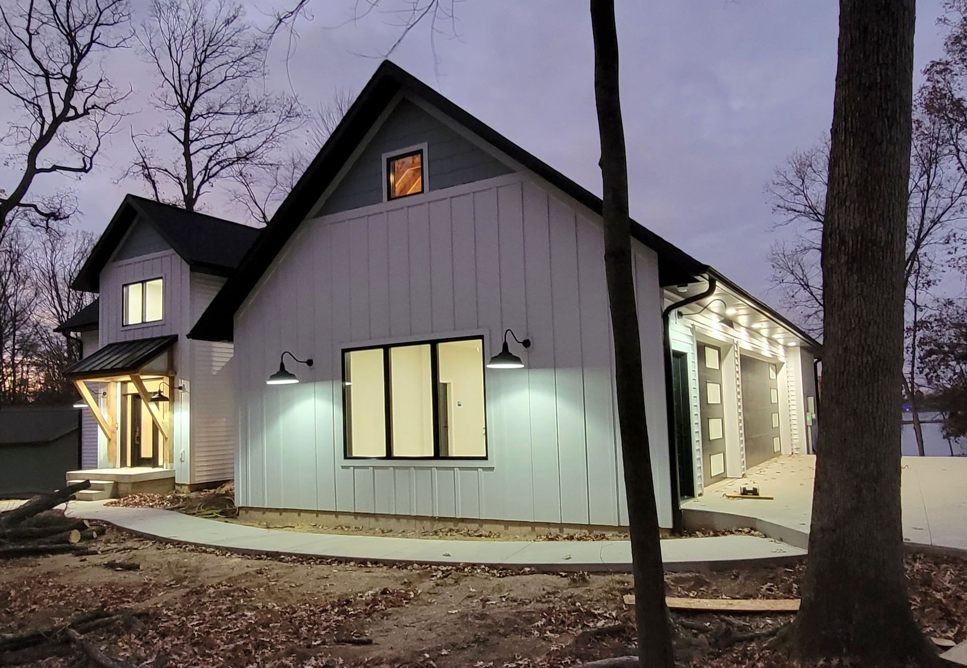 A white farmhouse-style home with black trim and a metal roof, situated in a wooded area at dusk.