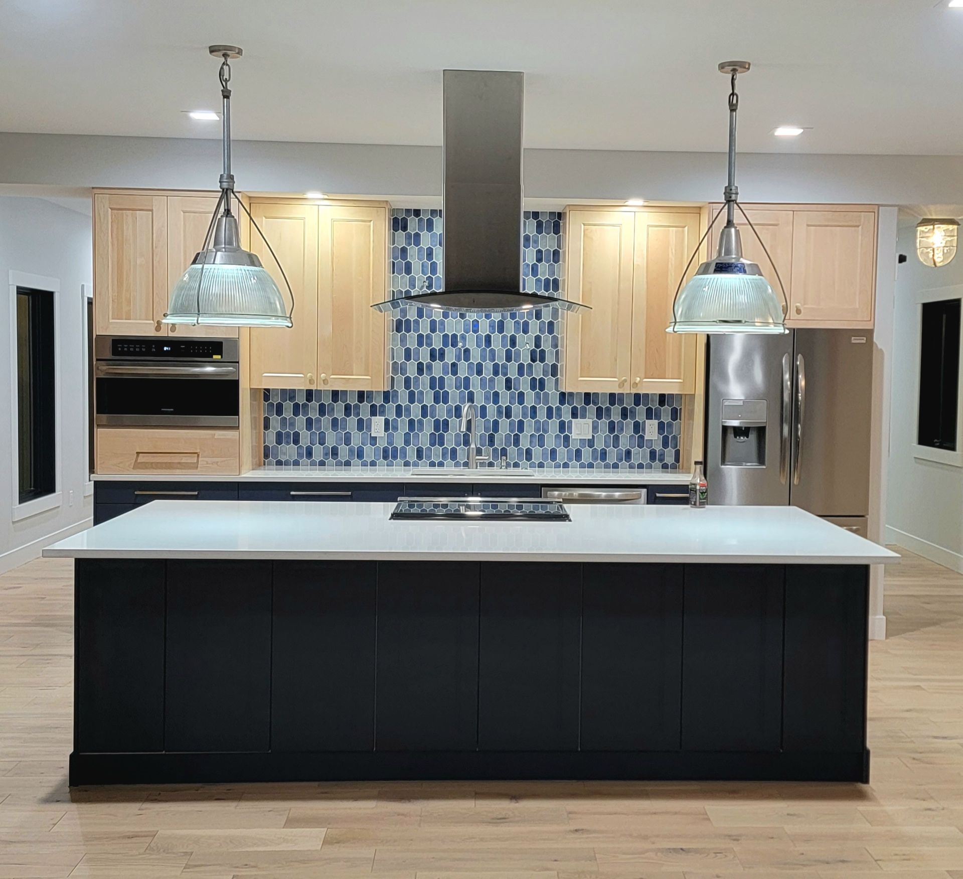A kitchen with a large black island, light wood cabinetry, blue patterned backsplash, and two hanging glass pendant lights.