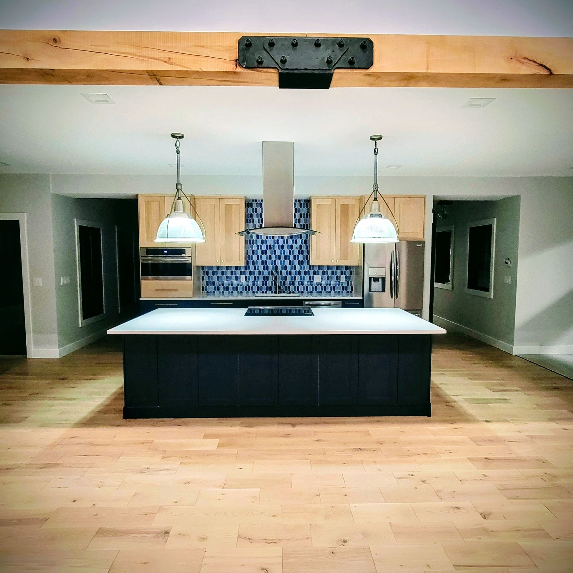 A spacious kitchen with a dark blue central island, light wood floors, two hanging pendant lights, and blue tile backsplash.