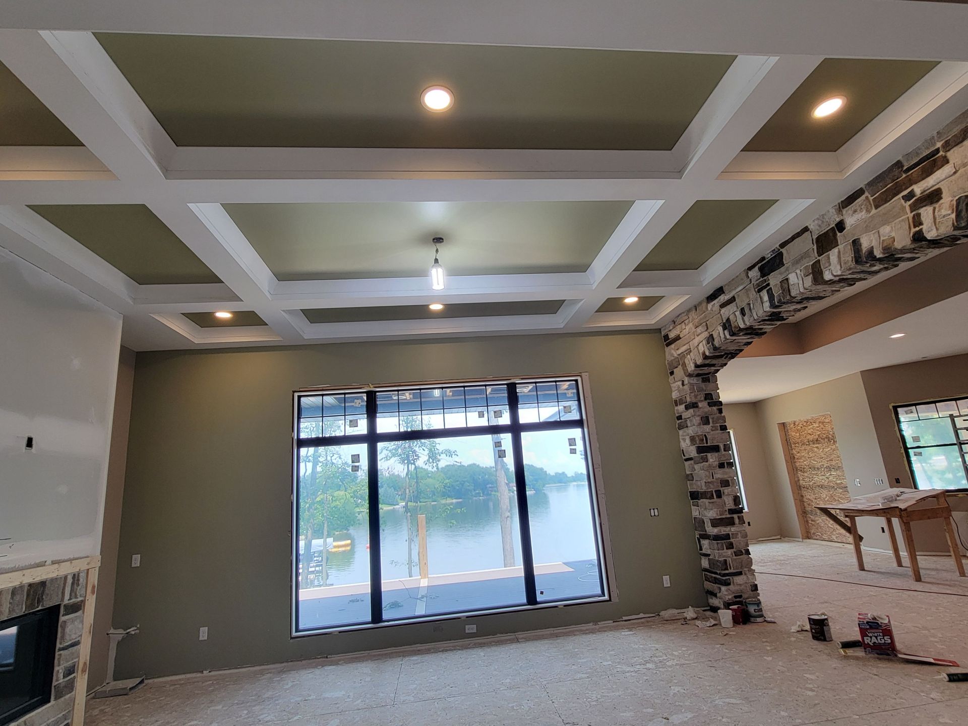 A room under construction with a coffered ceiling, green walls, a large window overlooking a lake, and a stone archway.