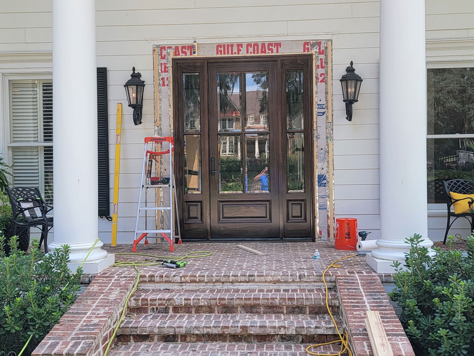 A brown wood front door with glass panels is being installed in a white home with brick steps and pillars.