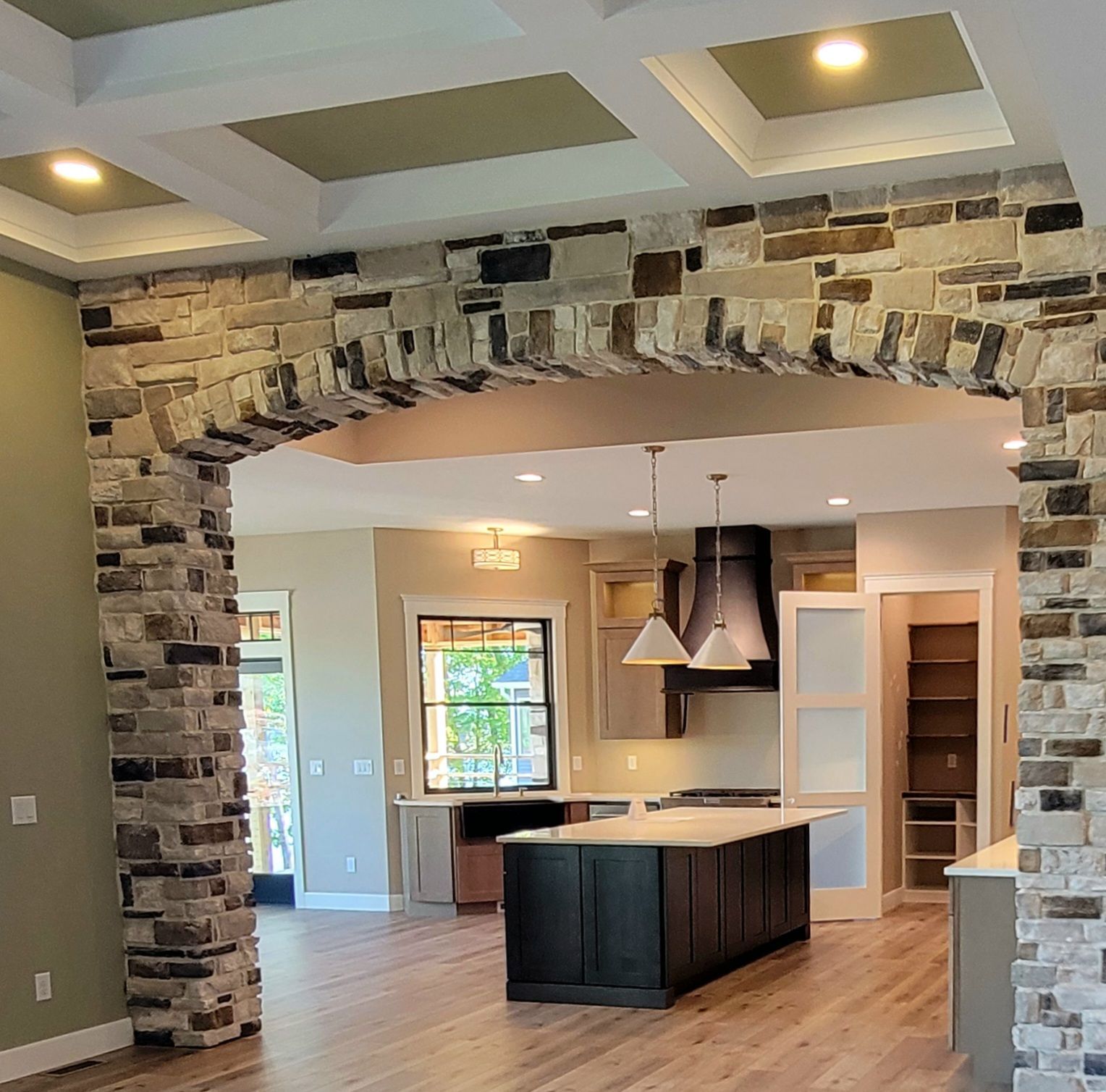 A rustic stone archway frames a modern kitchen featuring a dark island, pendant lighting, and light wood flooring.