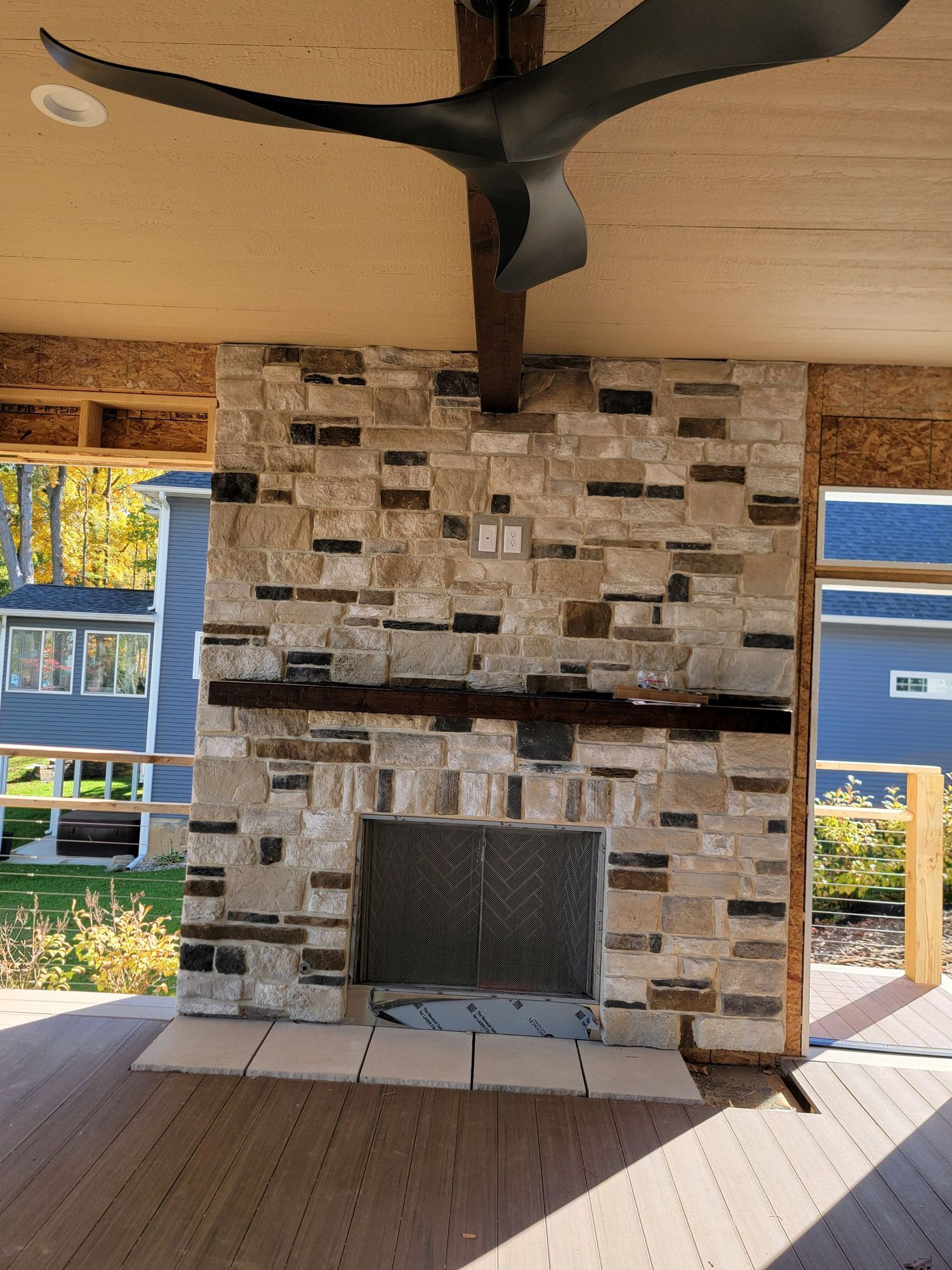 An outdoor stone fireplace with a dark wooden mantel and ceiling fan on a covered deck.