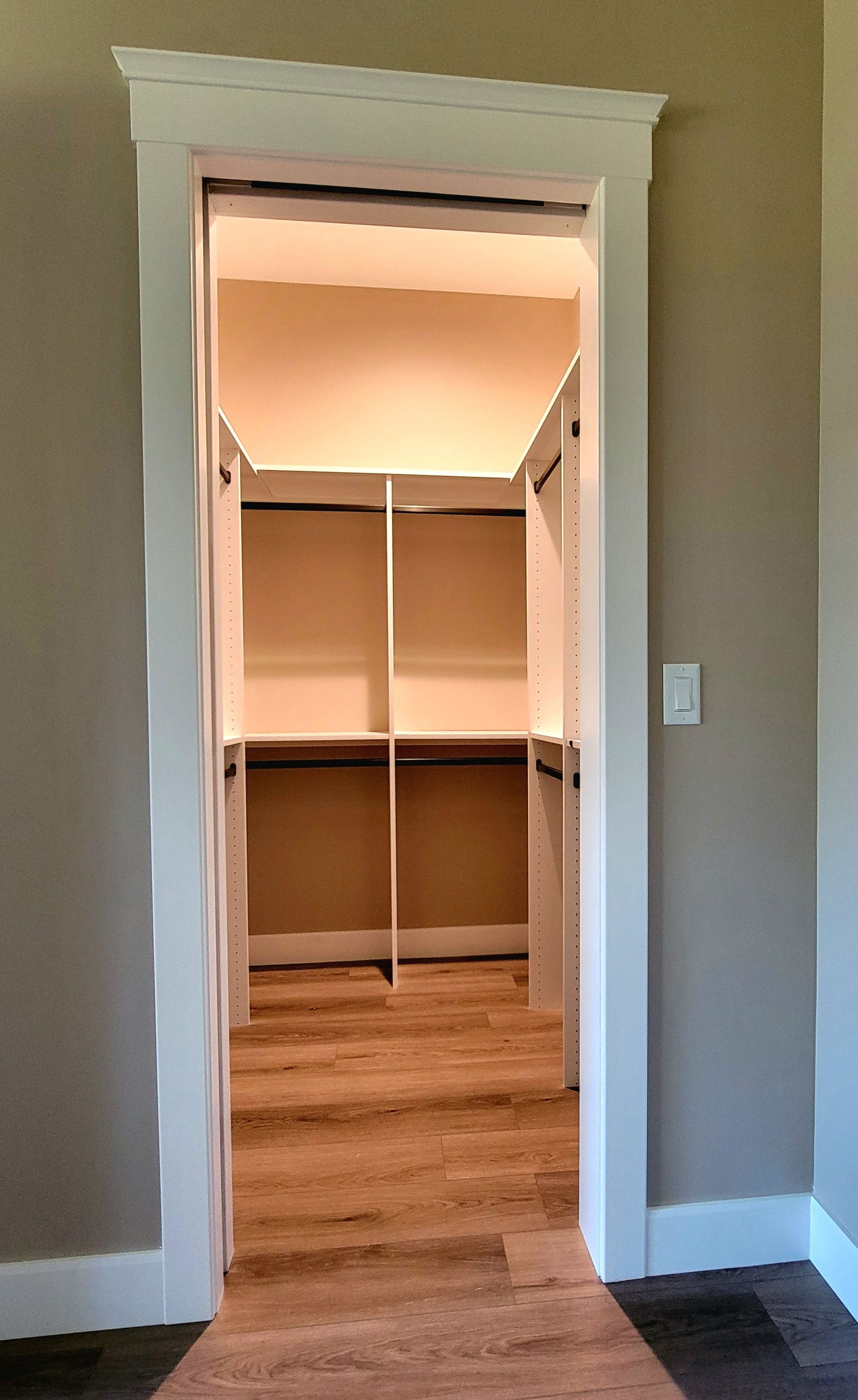 A walk-in closet viewed through a doorway with white trim, featuring white shelving and wood-tone flooring.