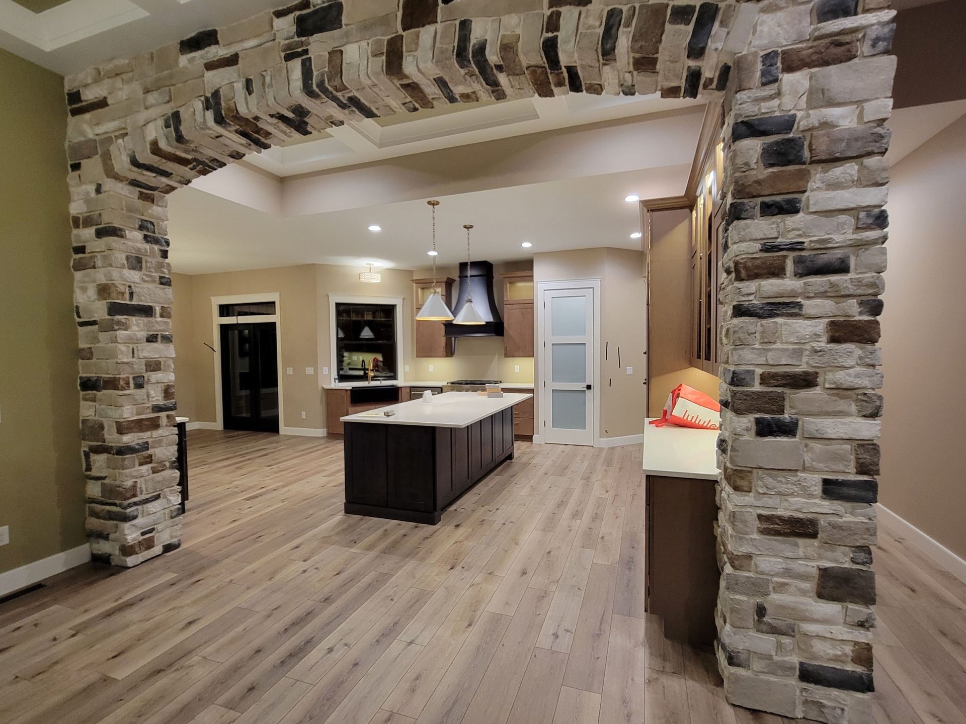 A kitchen view framed by a stone archway, featuring a dark central island, light wood floors, and neutral-toned cabinetry.