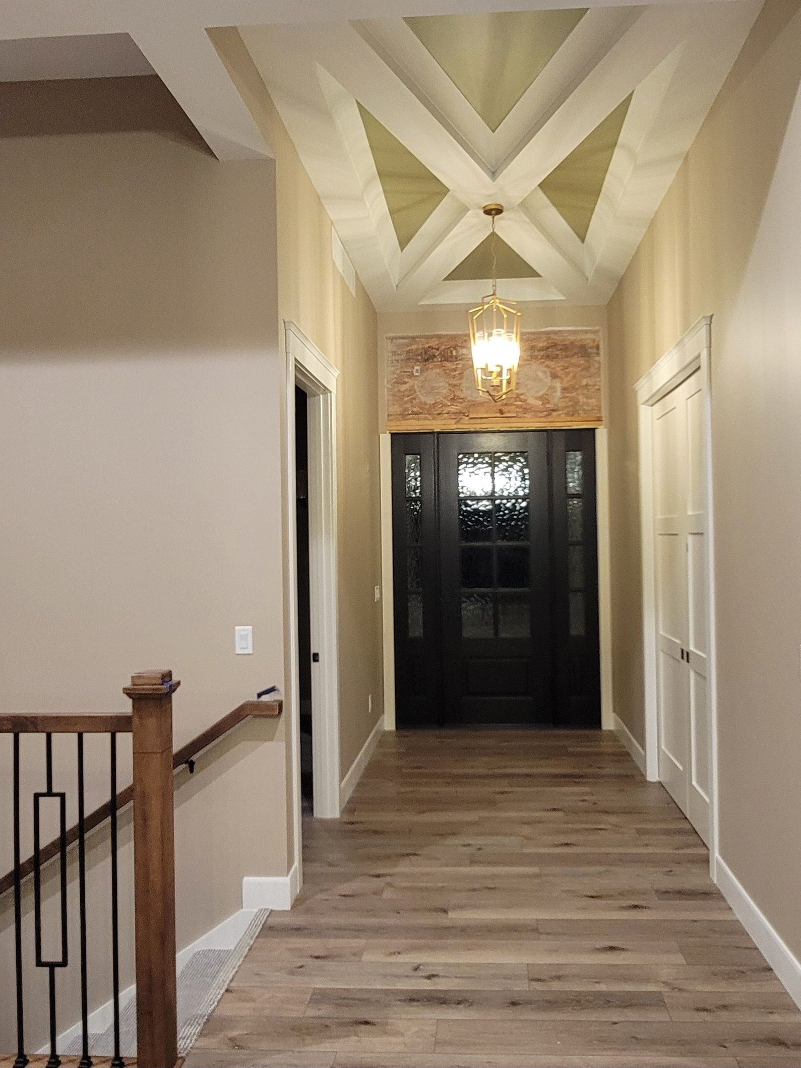 A hallway with light wood floors, a geometric patterned ceiling, a gold chandelier, and a dark front door with glass.