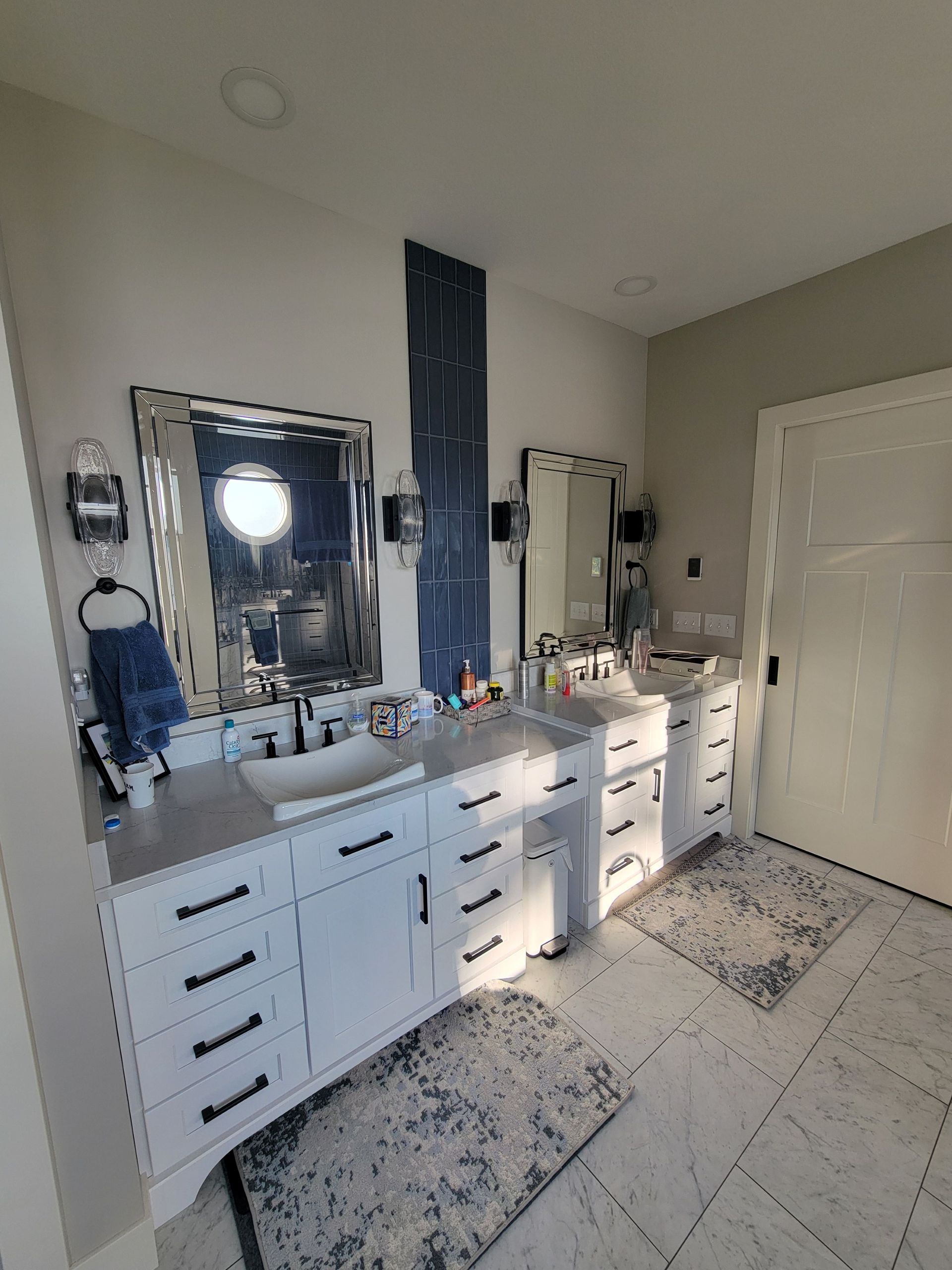 A double-sink bathroom vanity with white cabinets, dark hardware, patterned rugs, and two mirrors on a grey wall.