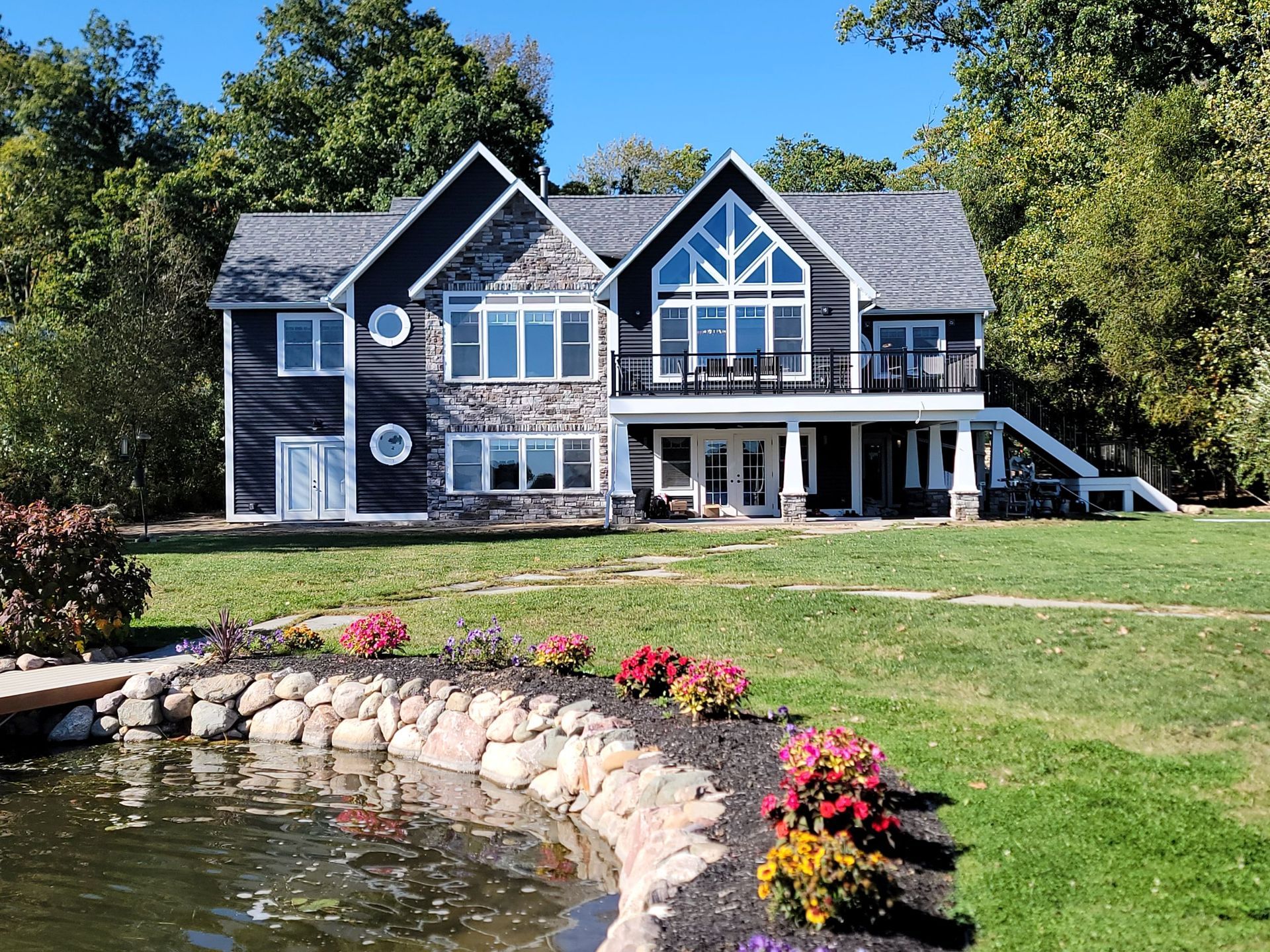 A two-story home with dark siding and stone accents sits behind a small pond with a stone-lined edge and landscaping.