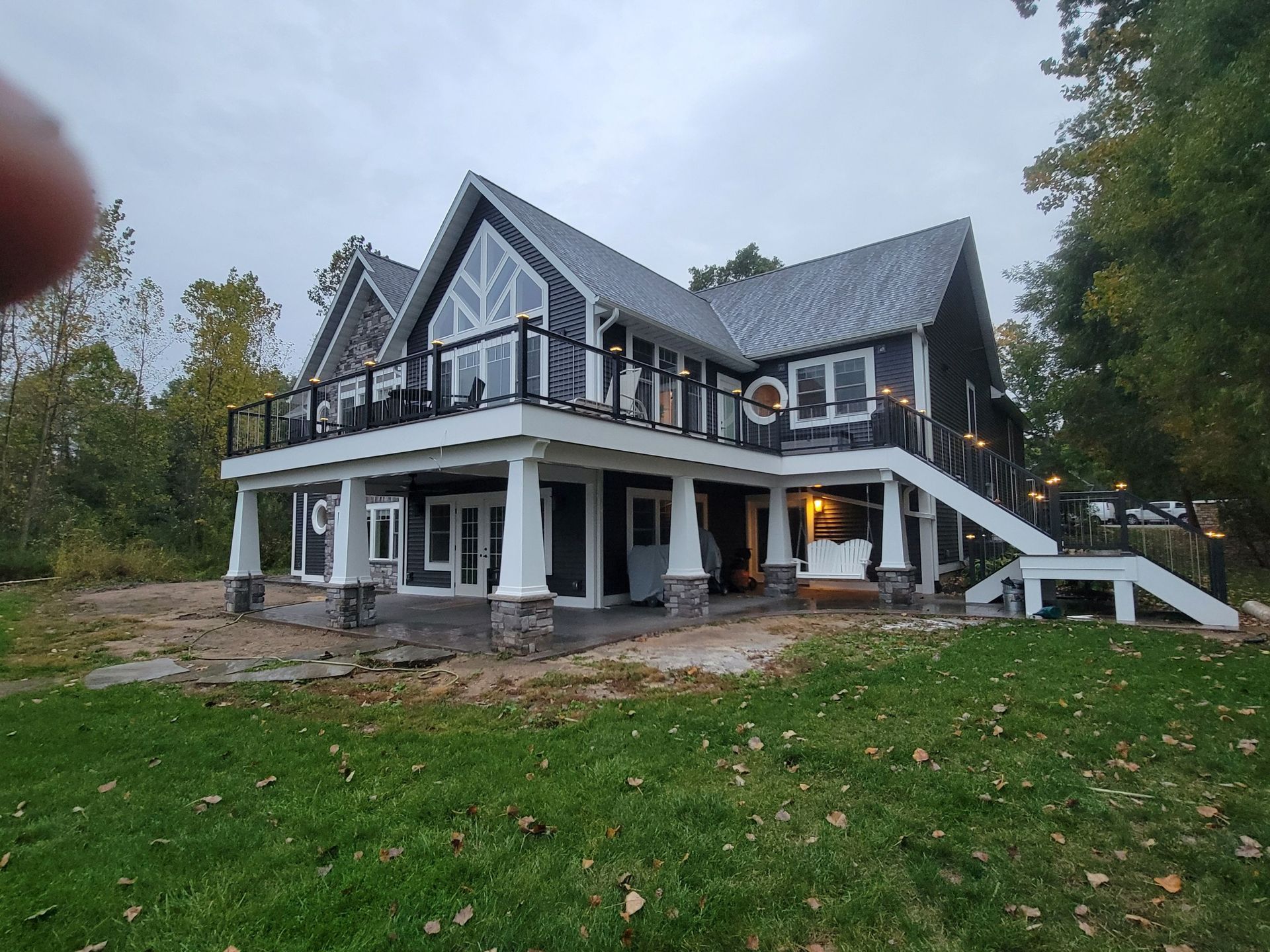 A two-story dark-sided house with a large elevated deck, white support columns, and a staircase leading to a grassy yard.