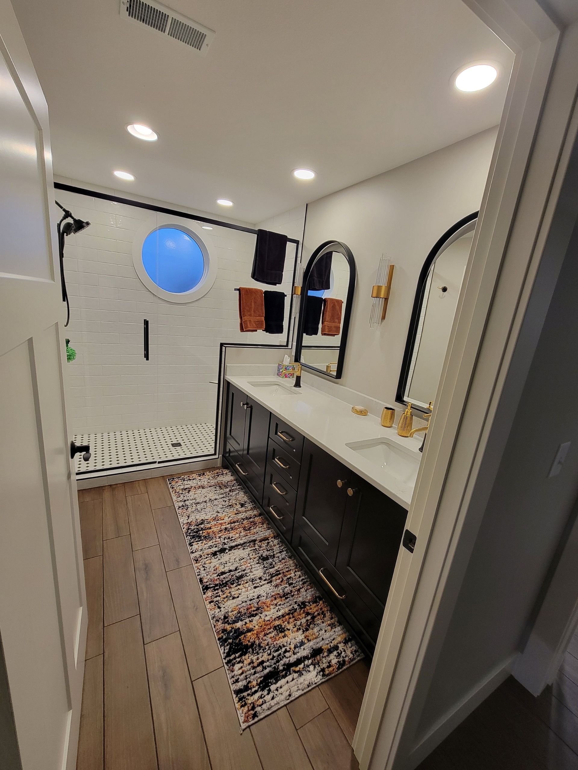 Modern bathroom with black cabinets, white countertops, two arched mirrors, wood-look tile flooring, and a round window.