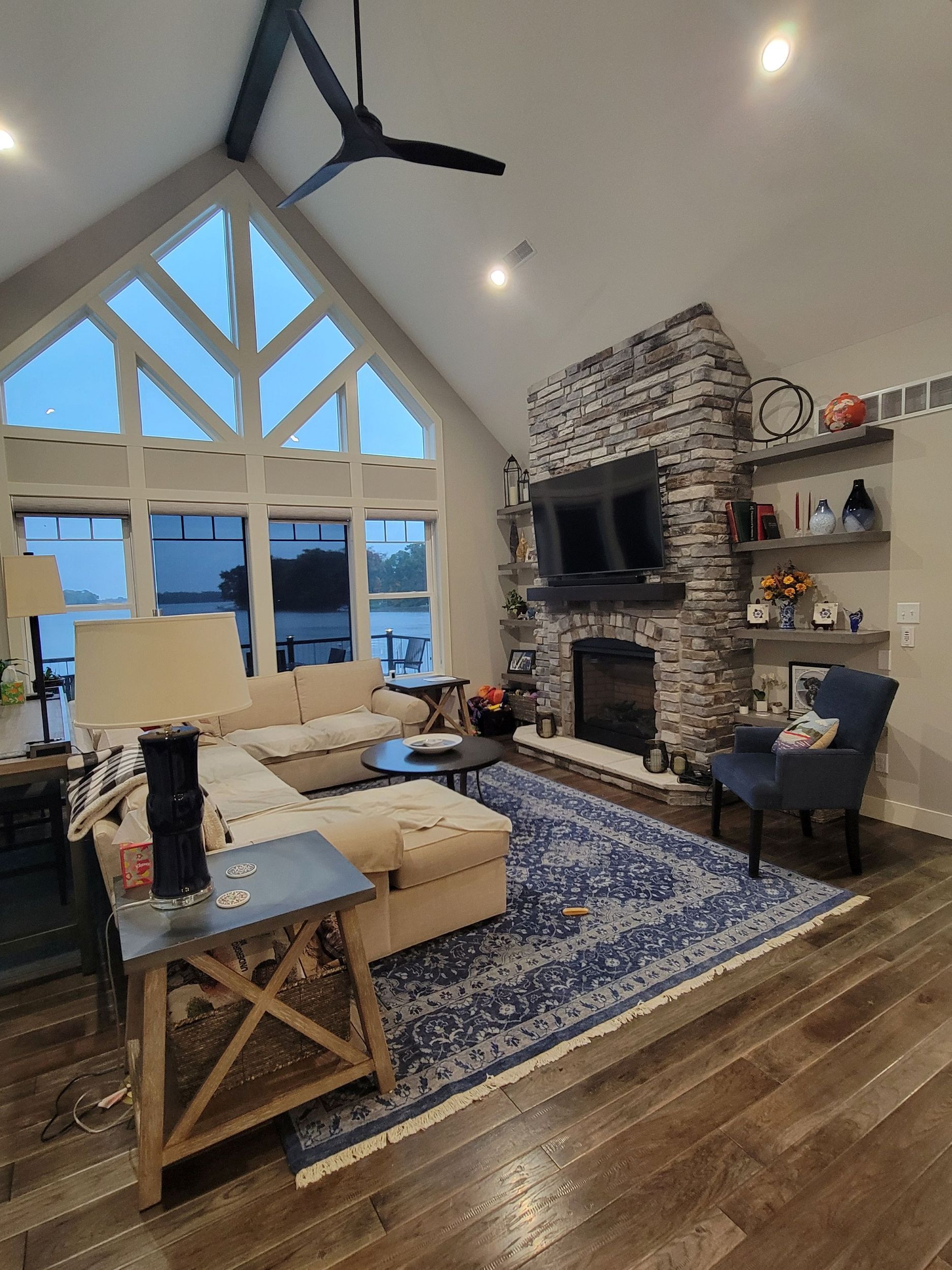 A bright living room with a large window wall, a stone fireplace, a blue patterned rug, and beige furniture.