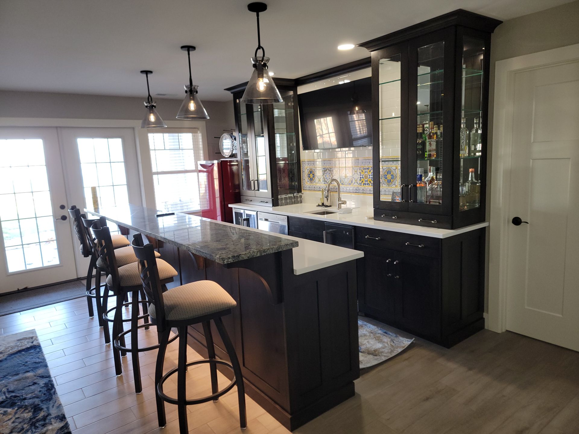 A modern home bar with dark cabinets, a granite-topped island, three stools, and glass-front display shelving.