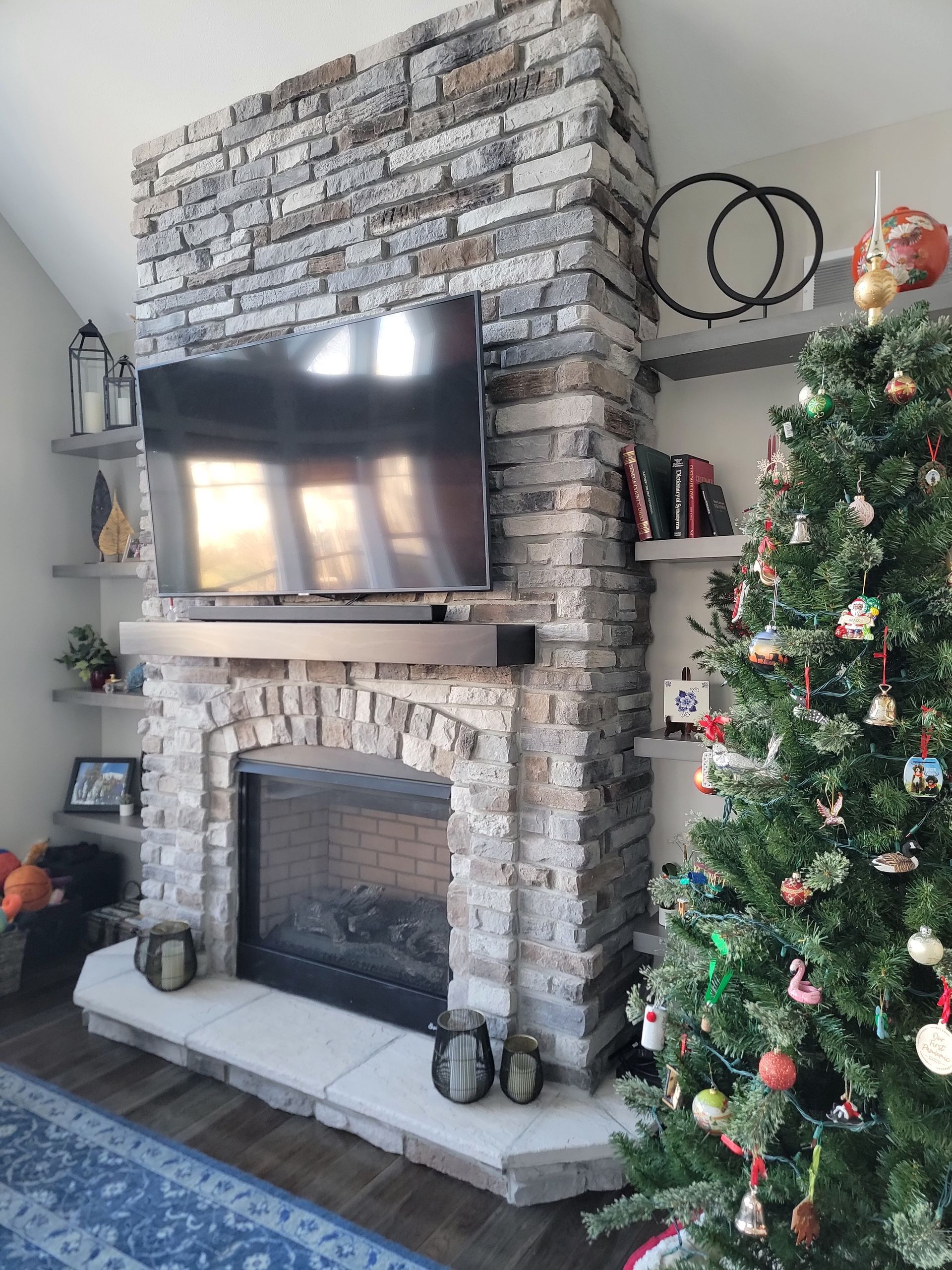 Stone fireplace with a mounted television above the mantle, flanked by shelves and a decorated Christmas tree.