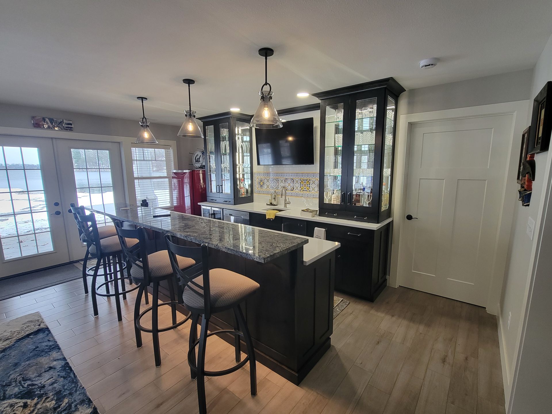 A home bar area with dark cabinetry, granite countertops, three bar stools, pendant lighting, and a small refrigerator.