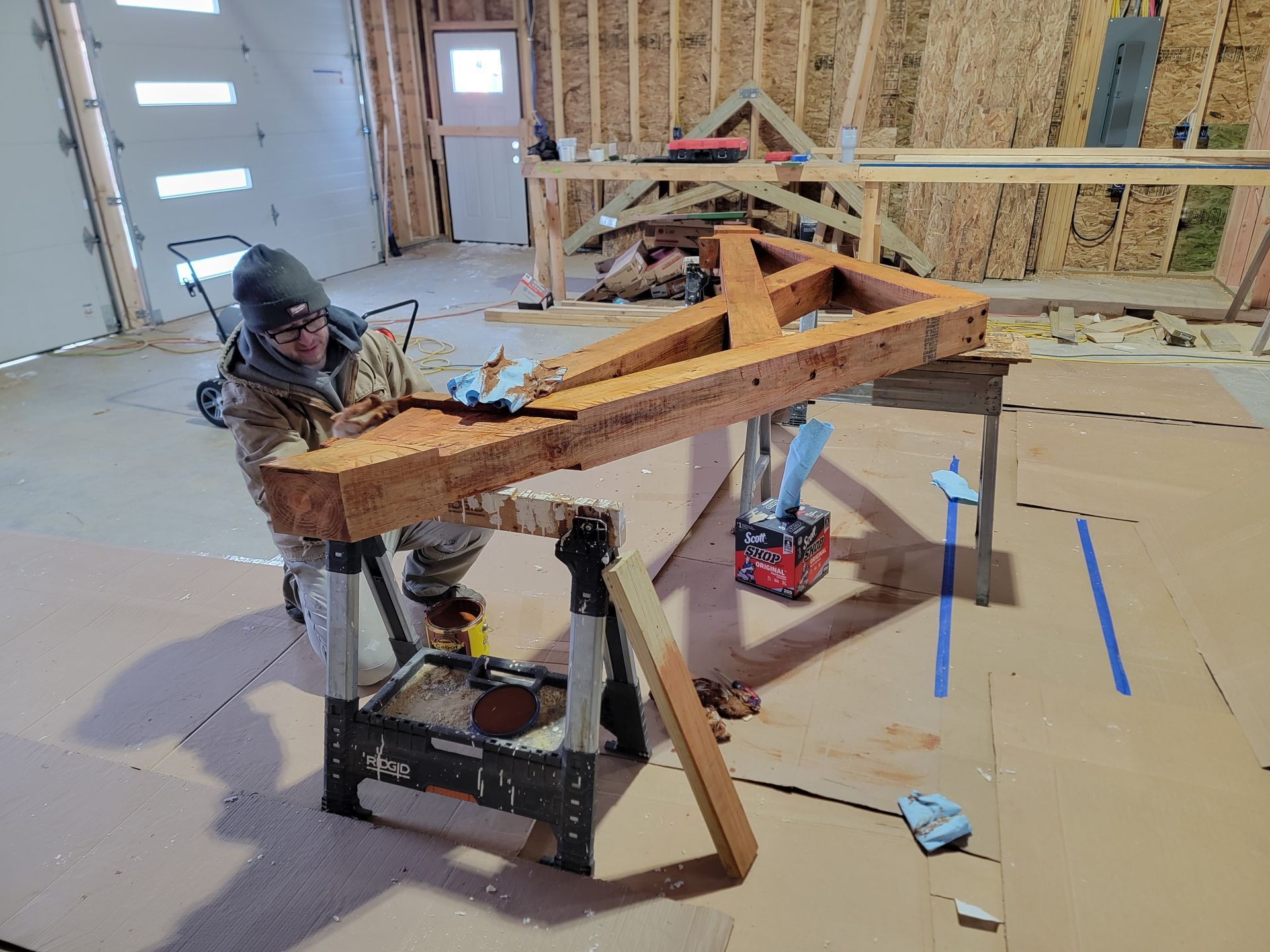 A person in a workshop applying stain to a large, wooden triangular truss frame supported by sawhorses.
