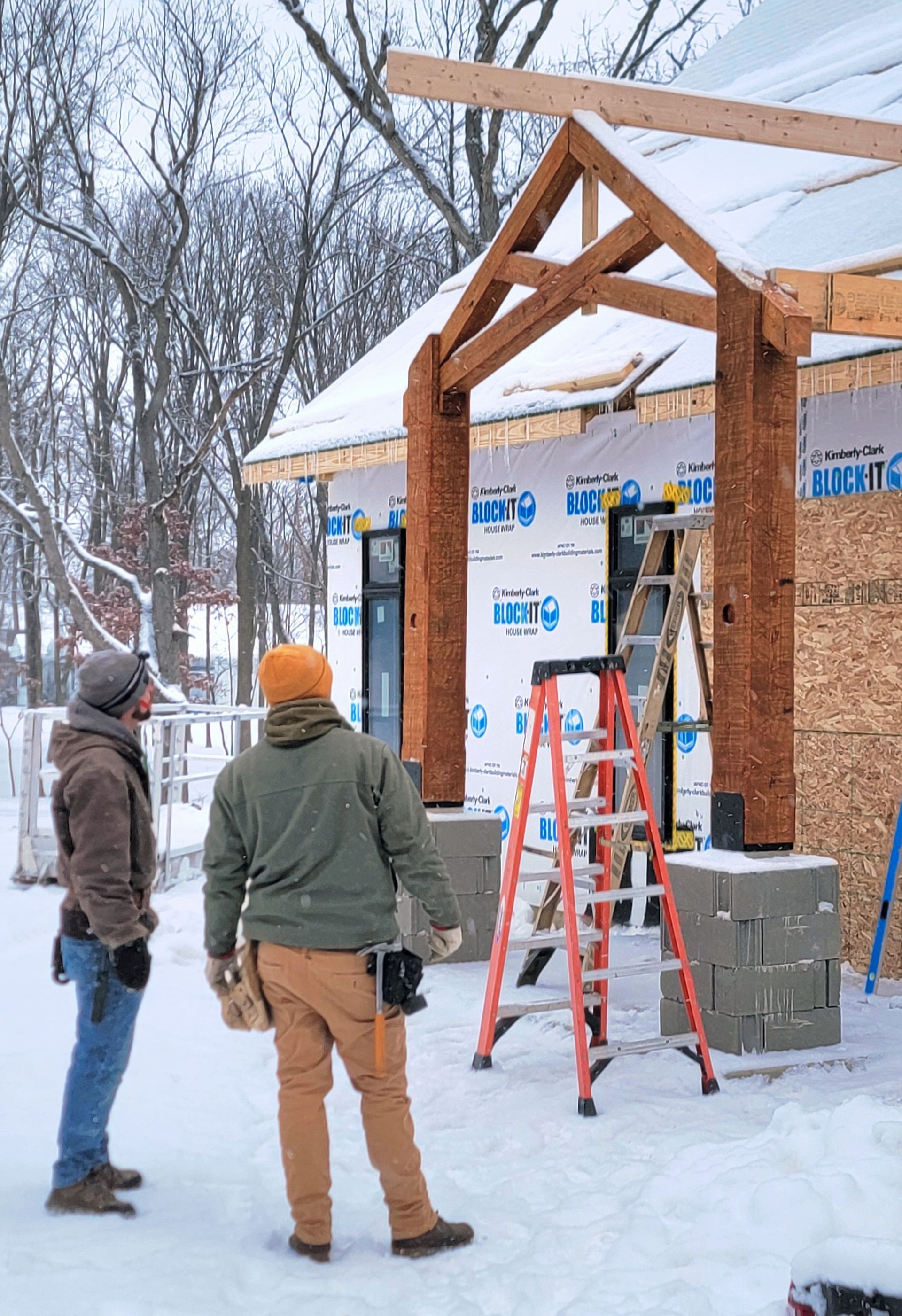 Two people stand in the snow looking at a wooden porch frame being built on the front of a house under construction.