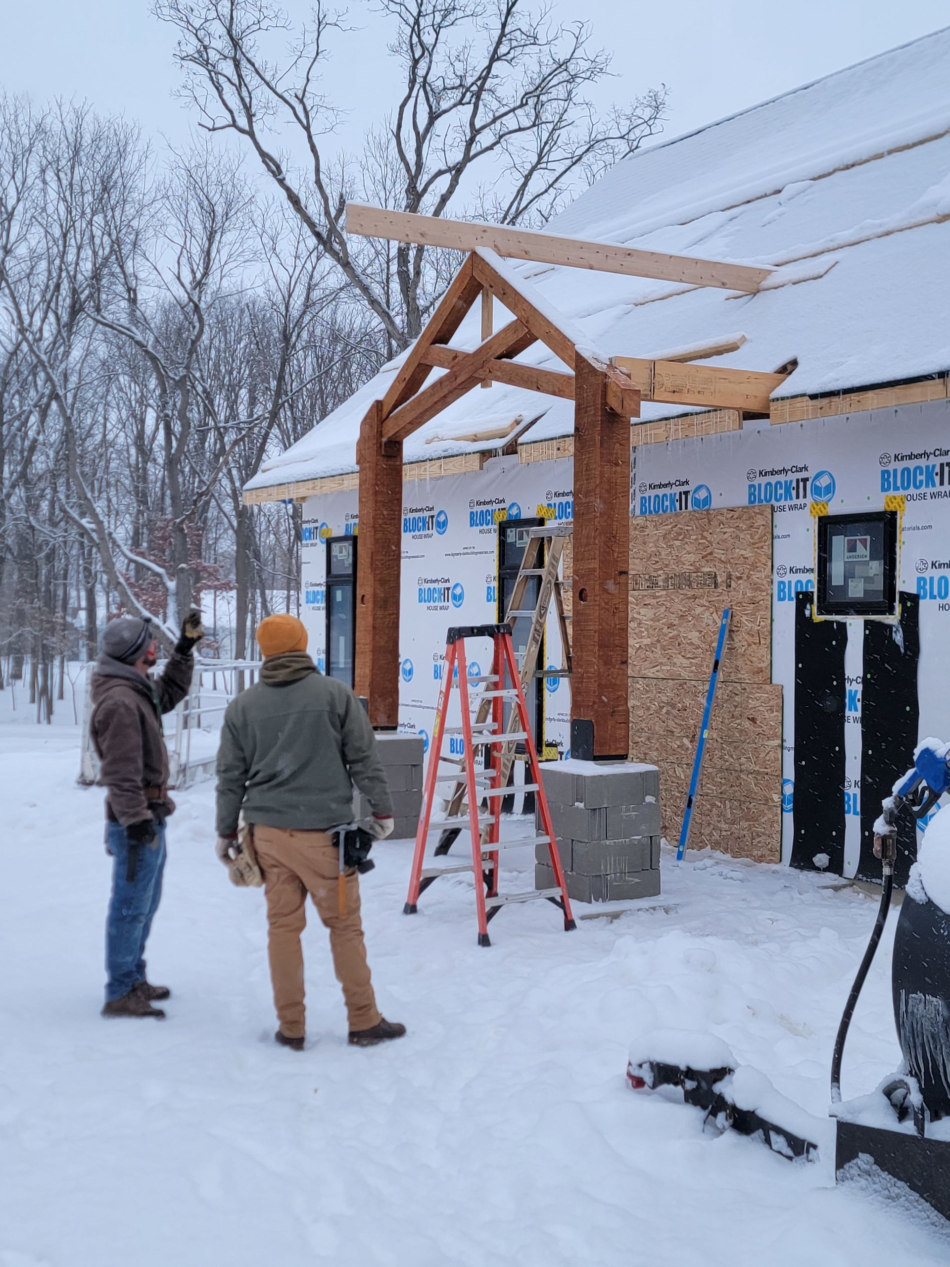 Two people stand in the snow outside a construction site, looking up at a partially finished wooden entryway structure.