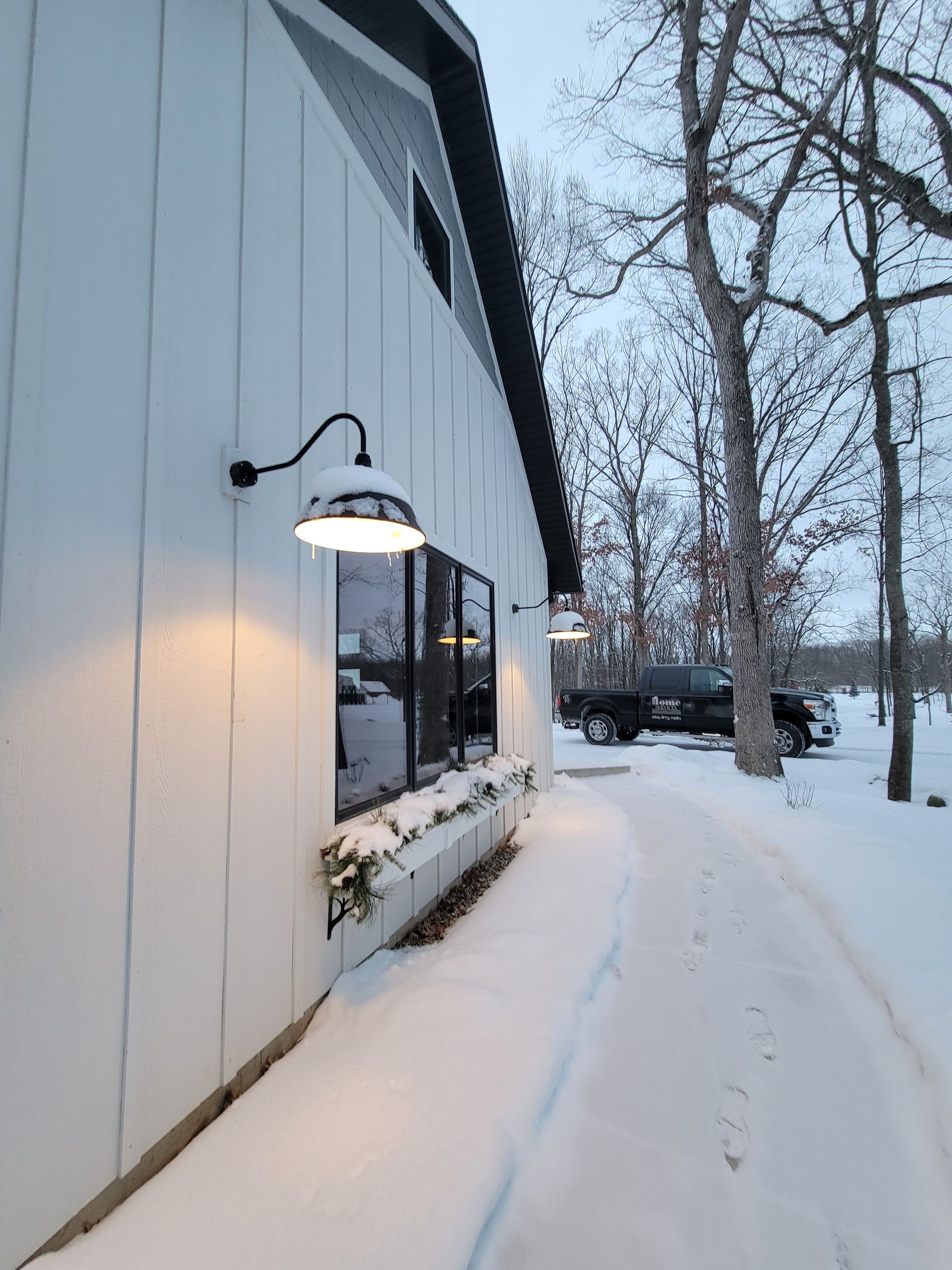A glowing outdoor barn-style light mounted on a white vertical-paneled wall, illuminating a snow-covered path and yard.
