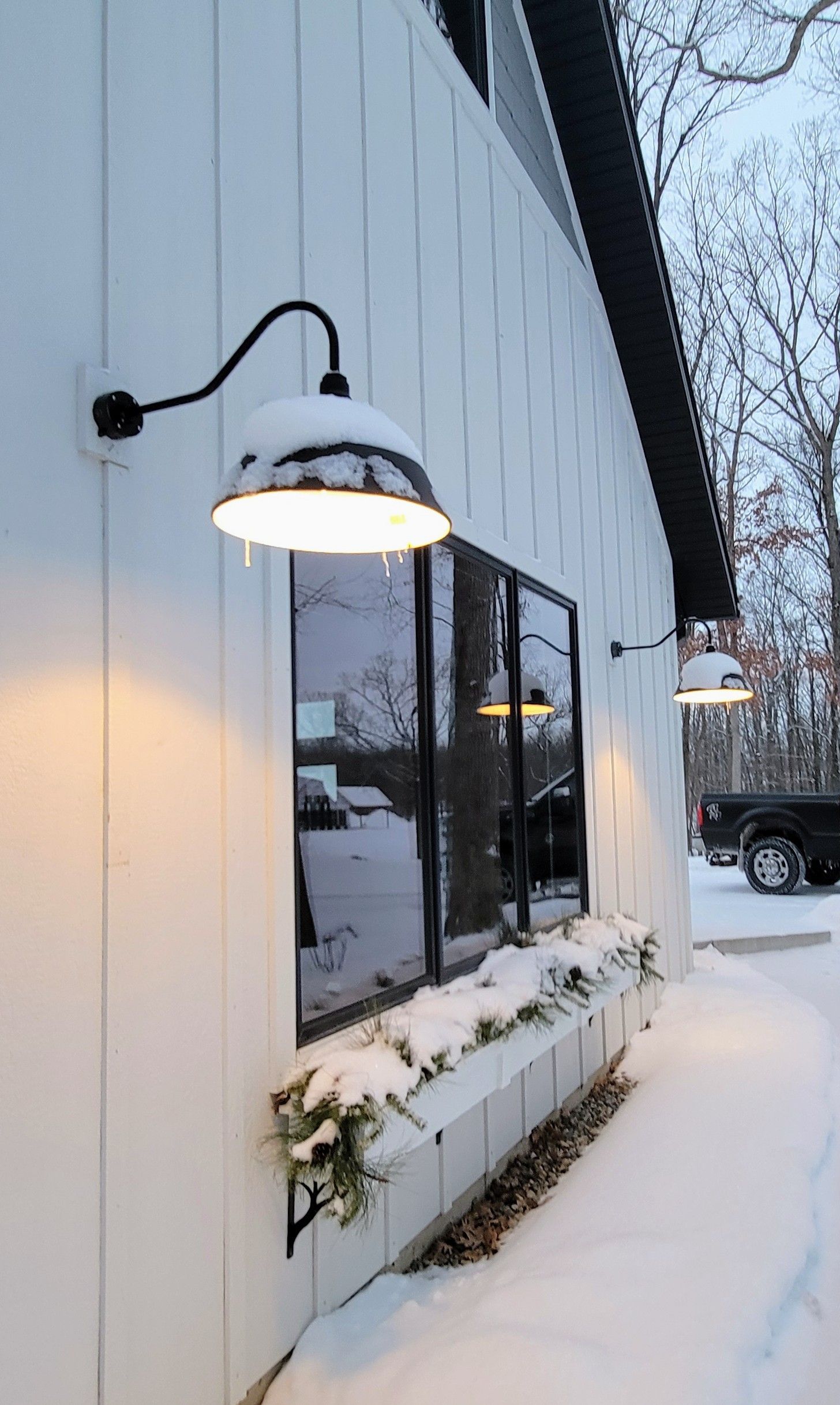 Exterior wall with board-and-batten siding and a window box, both covered in snow, lit by two outdoor gooseneck lamps.