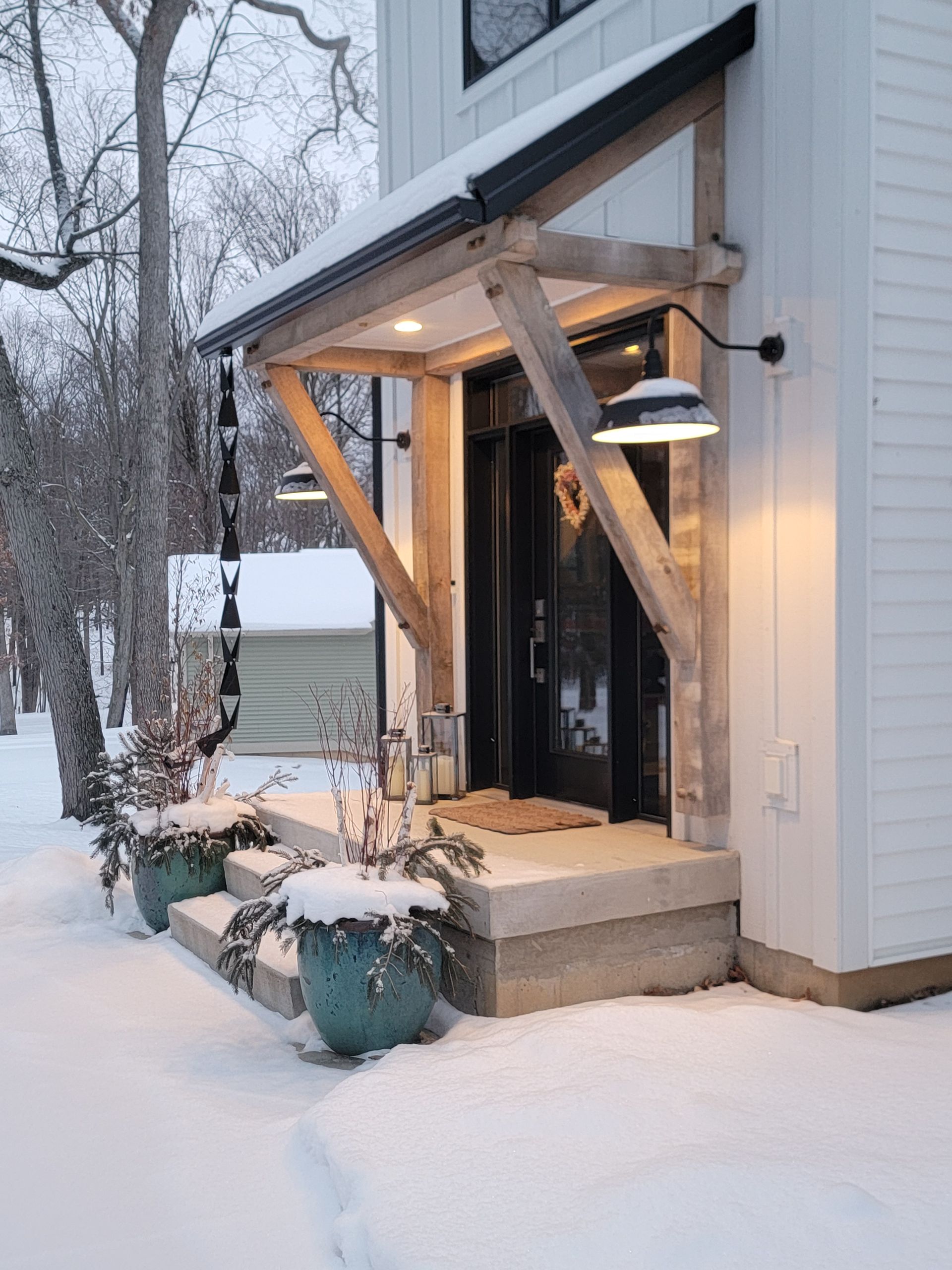 A modern white farmhouse entryway with a timber-frame porch, black lighting, and two teal planters filled with snow.