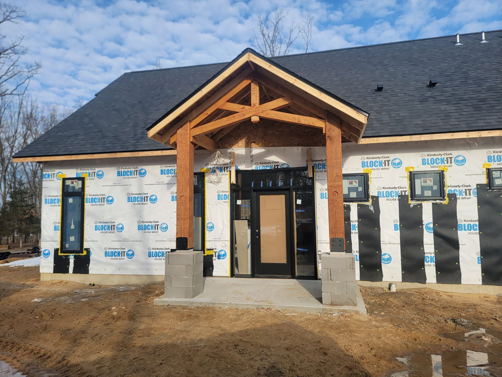A house under construction with a timber-framed porch, dark roof shingles, and unfinished exterior wall sheathing.