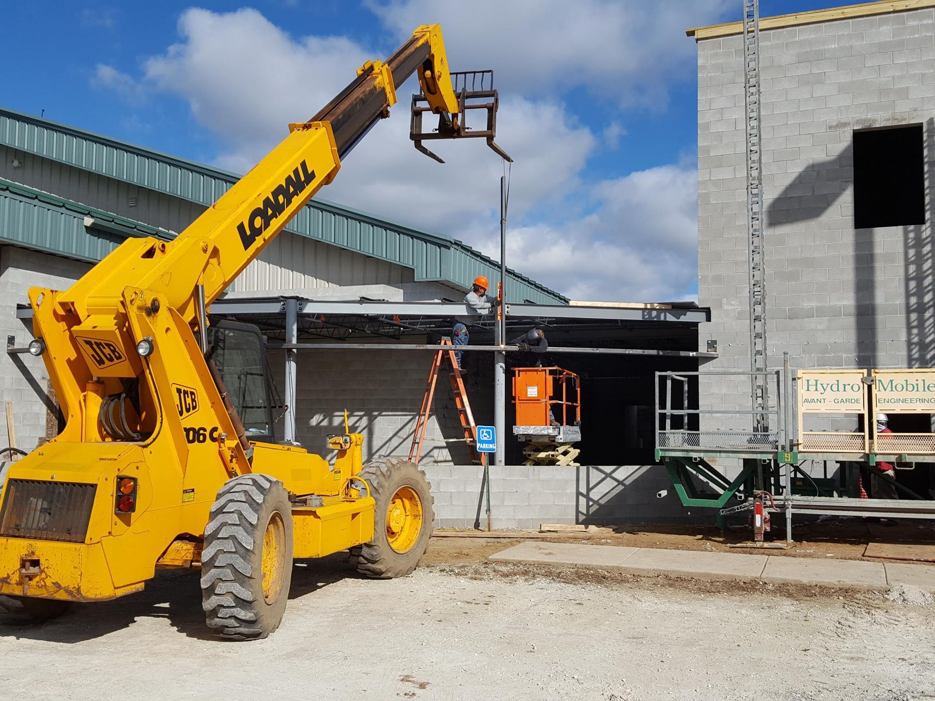 A bright yellow telescopic handler sits at a construction site with workers near a block building under a cloudy sky.