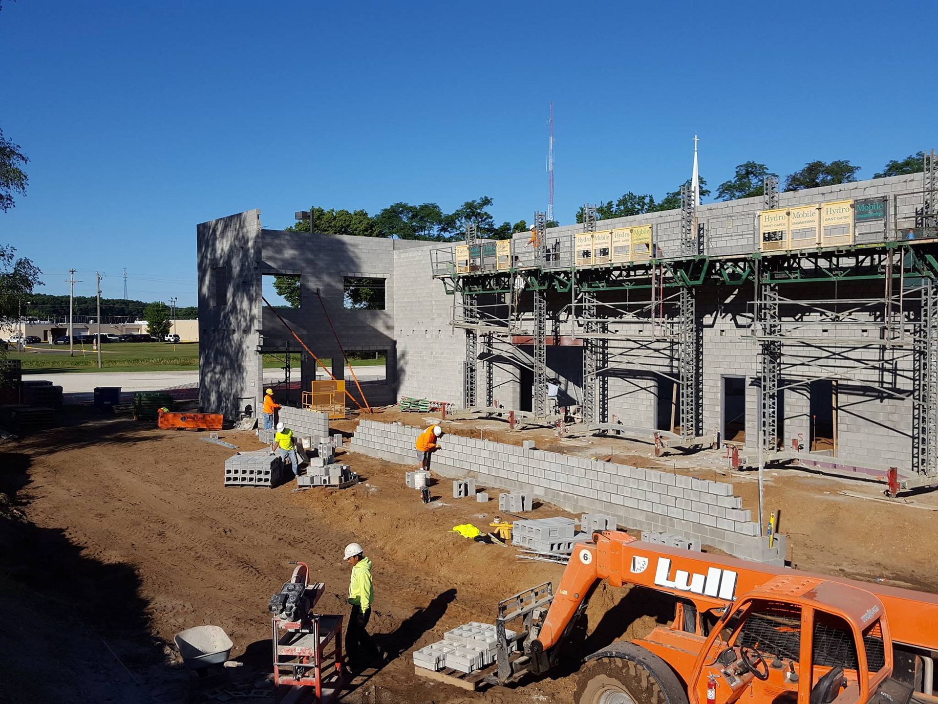 Construction workers laying concrete blocks at a building site with an orange telehandler in the foreground.