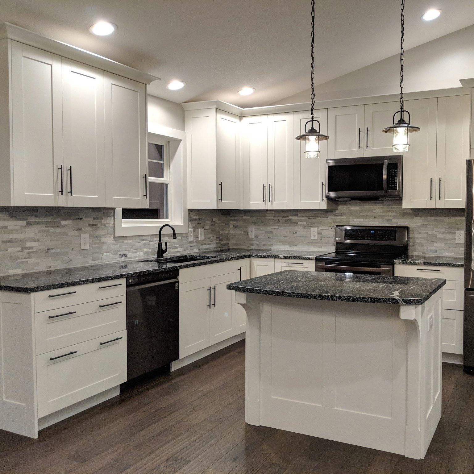 A white kitchen with dark countertops, wood floors, a small center island, and two pendant lights hanging above it.