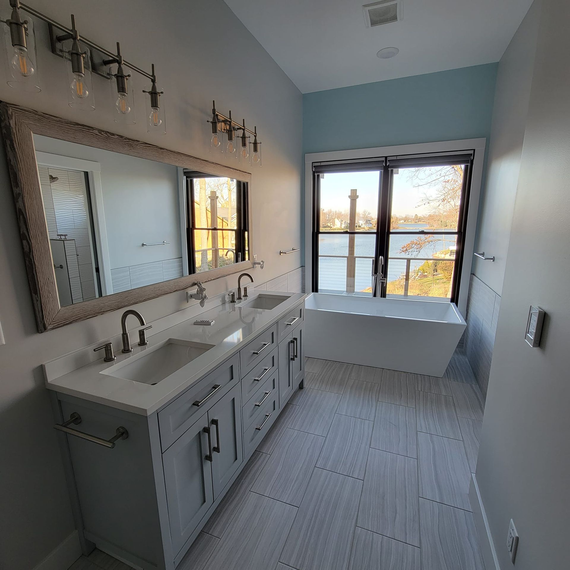 A modern bathroom with a double vanity, a large framed mirror, and a freestanding white bathtub by a window overlooking water.