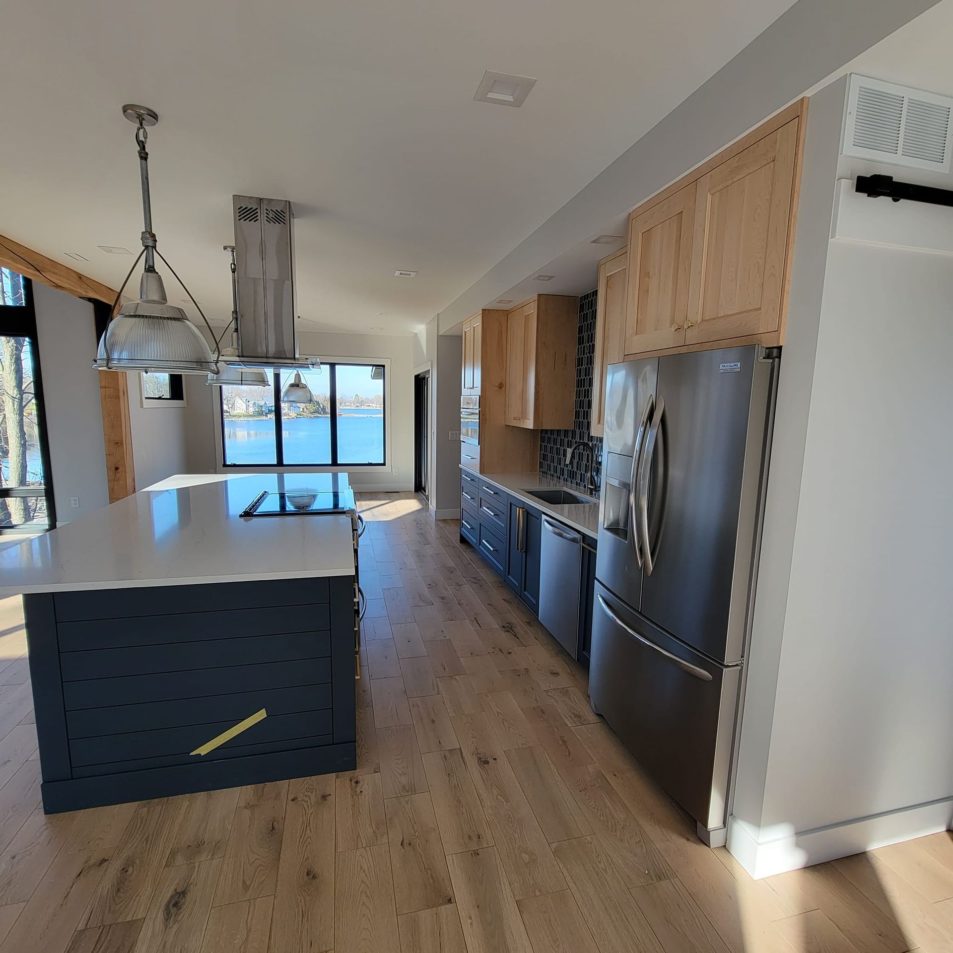 A modern kitchen with a dark blue island, light wood cabinets, stainless steel appliances, and a view of the water.