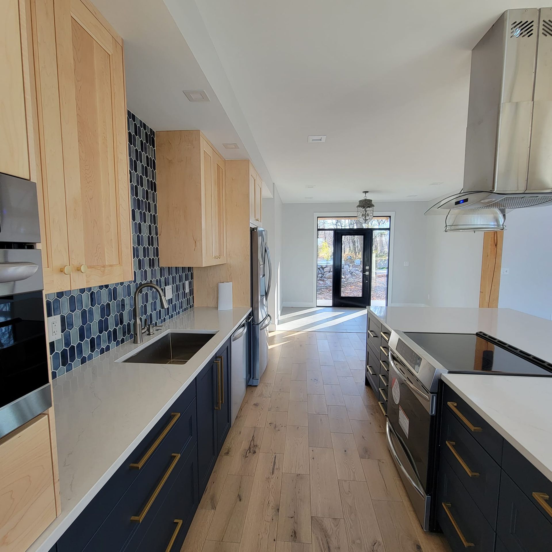 A bright kitchen featuring dark blue cabinets, light wood upper cabinets, white countertops, and a blue patterned backsplash.