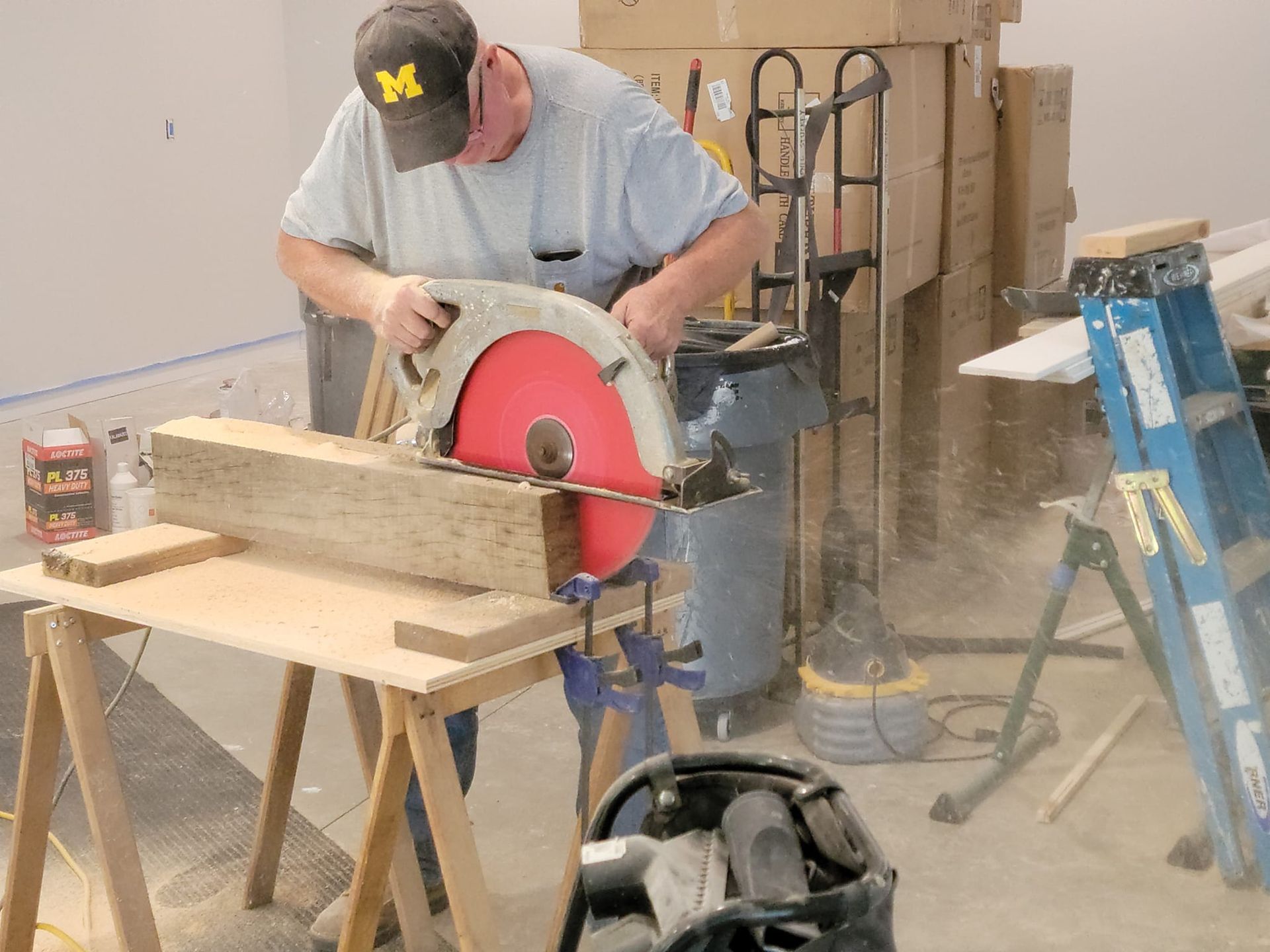 A person in a University of Michigan cap uses a circular saw to cut a large wooden beam on a workbench.