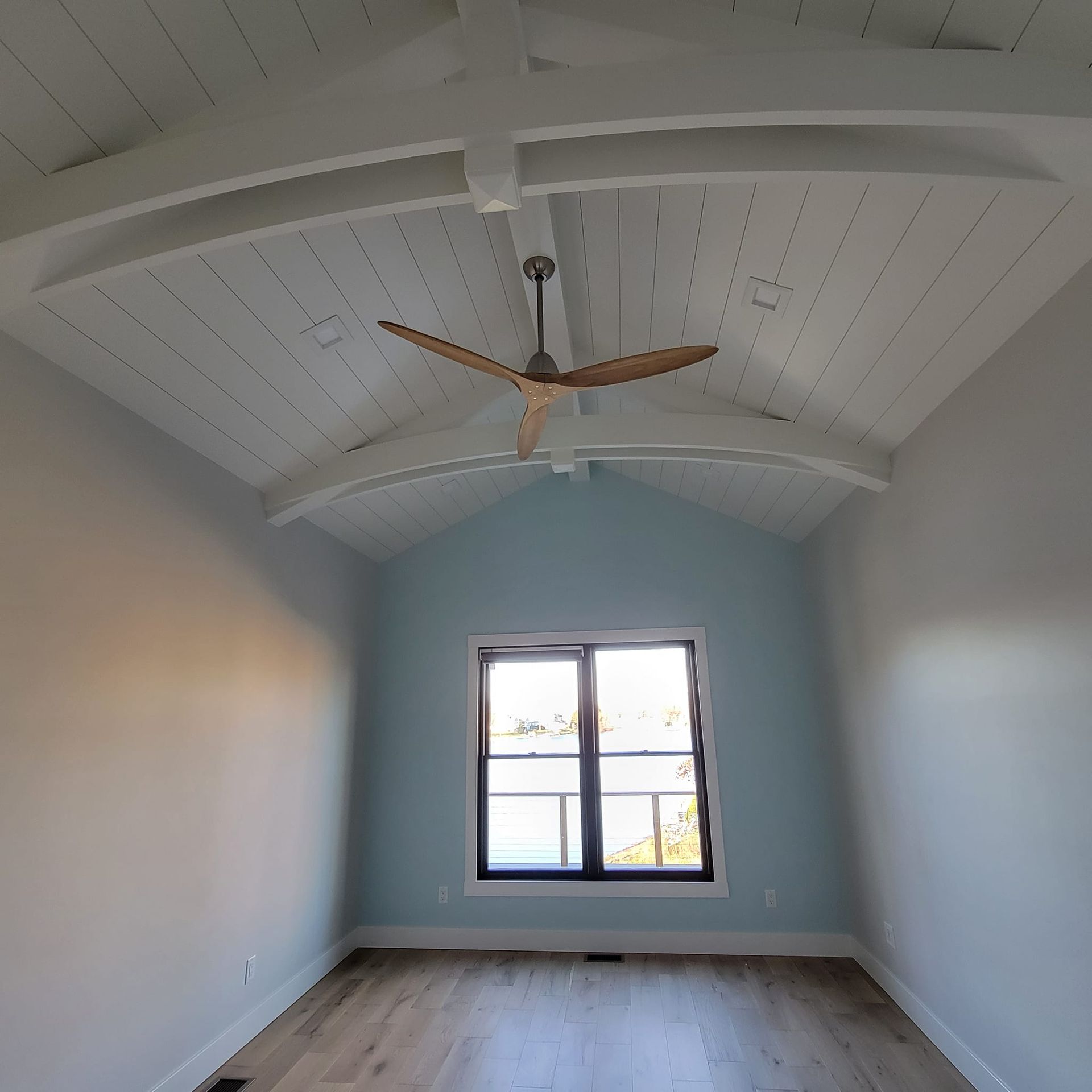 Empty room with a vaulted white-beamed ceiling, light wood floors, gray walls, and a light blue accent wall with a window.