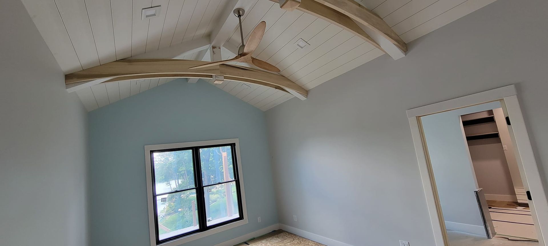 Empty bedroom with vaulted plank ceiling, exposed wooden beams, a ceiling fan, a window, and an open doorway to a closet.