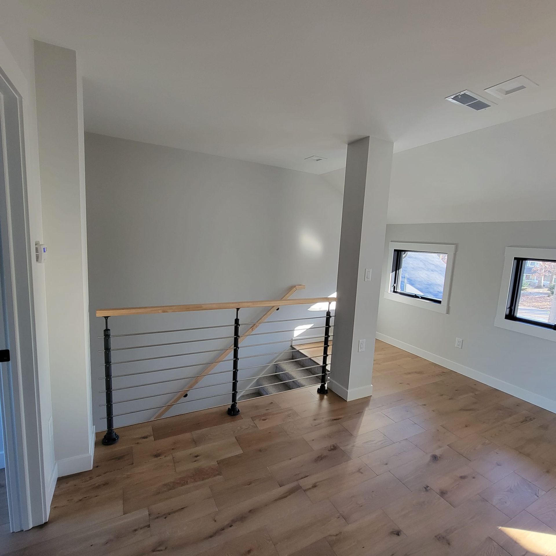 A modern loft landing with light wood floors, a cable railing staircase, and two windows on a light gray wall.