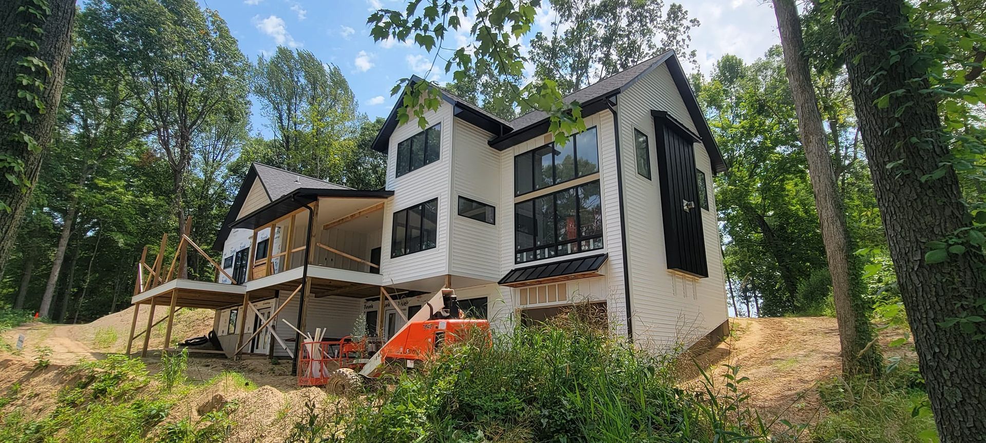 A two-story house under construction with white siding, large black-framed windows, and a wooden deck, set in a wooded area.