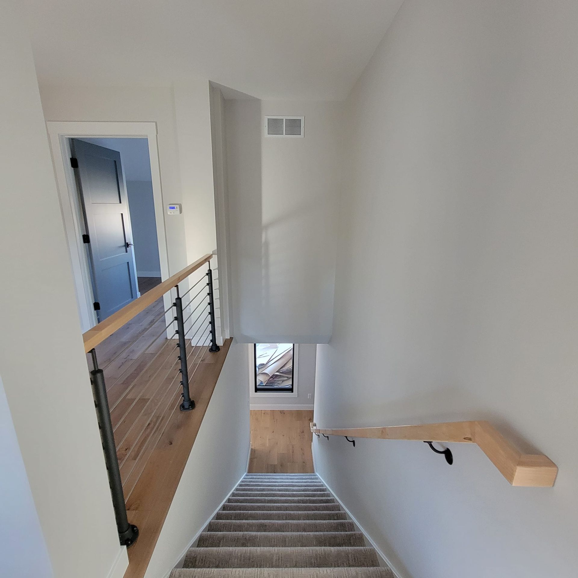 A view from the top of carpeted stairs looking down to a landing, with a wooden handrail and cable railing to the side.
