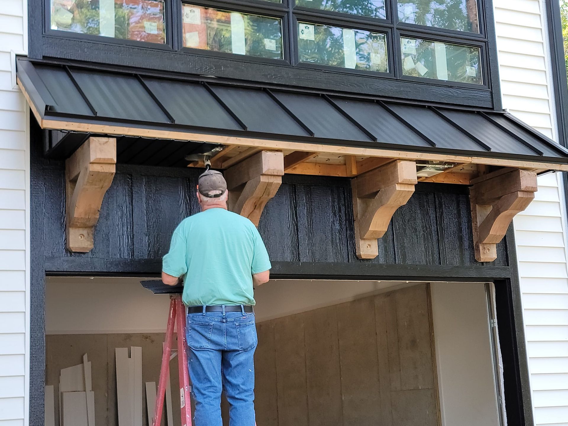 A person on a ladder installs an unfinished wooden bracket under the black metal awning of a garage doorway.