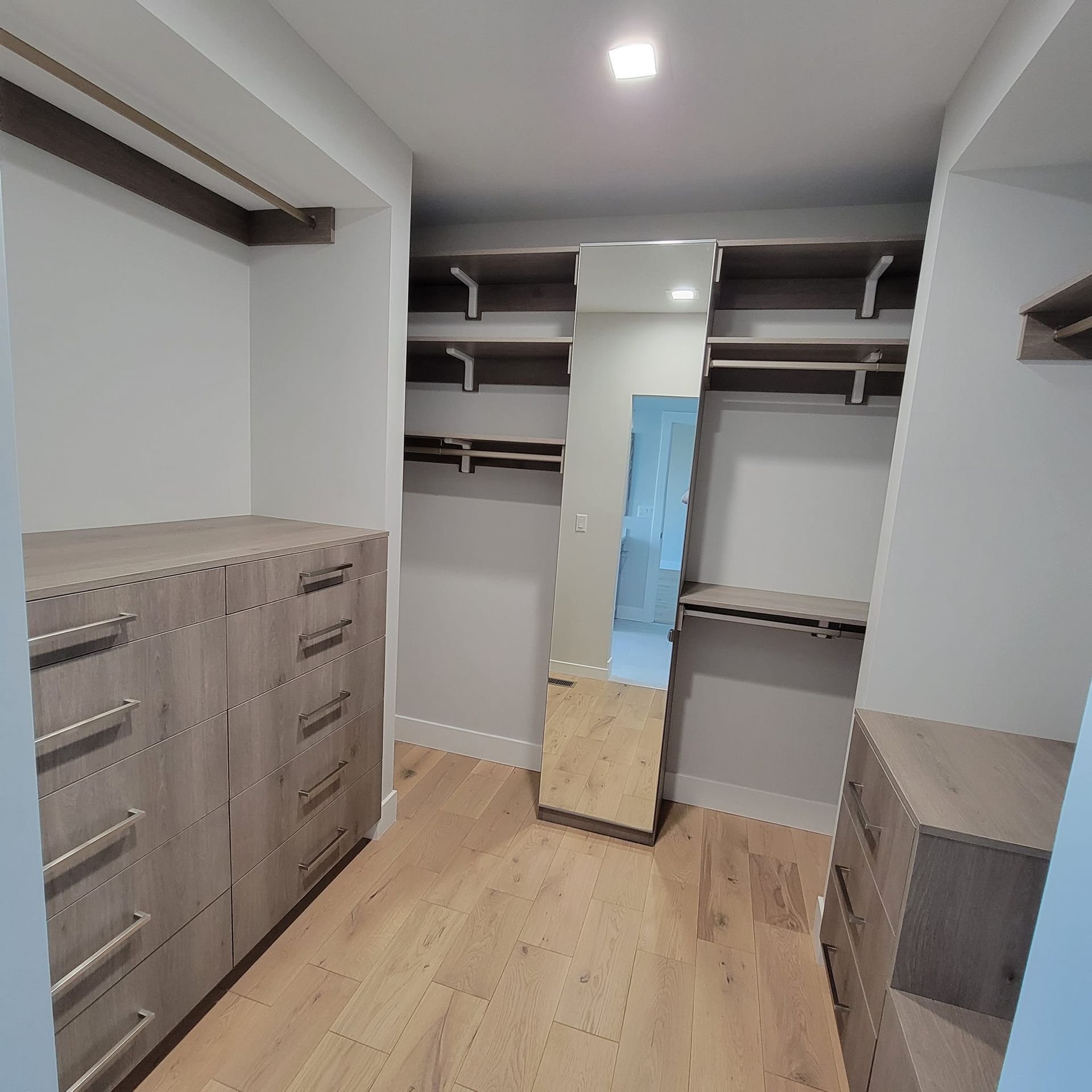 A walk-in closet featuring light wood-grain cabinetry, built-in shelving, a full-length mirror, and light hardwood floors.